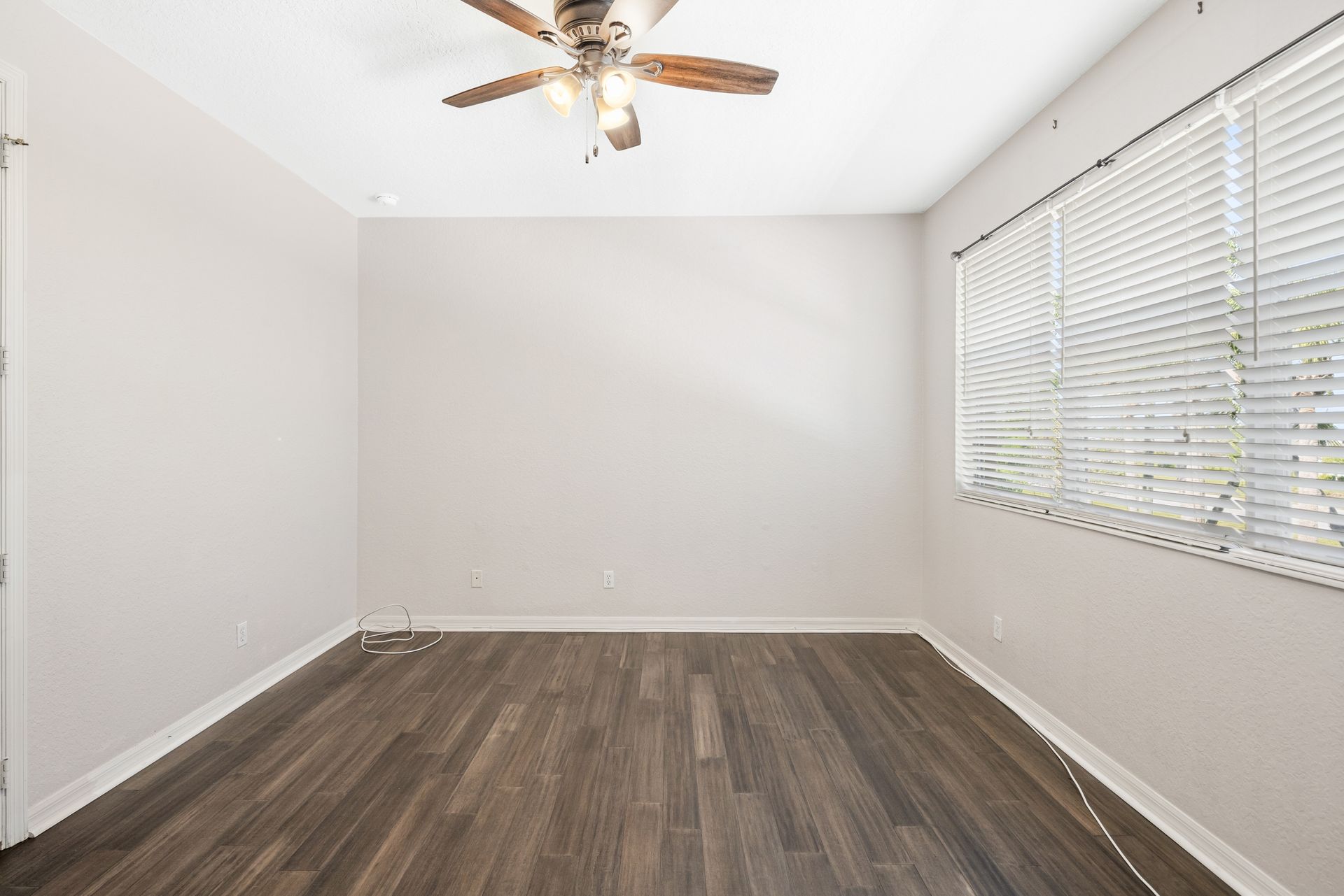 An empty room with light gray walls, dark wood-style flooring, a ceiling fan, and a window with white horizontal blinds.
