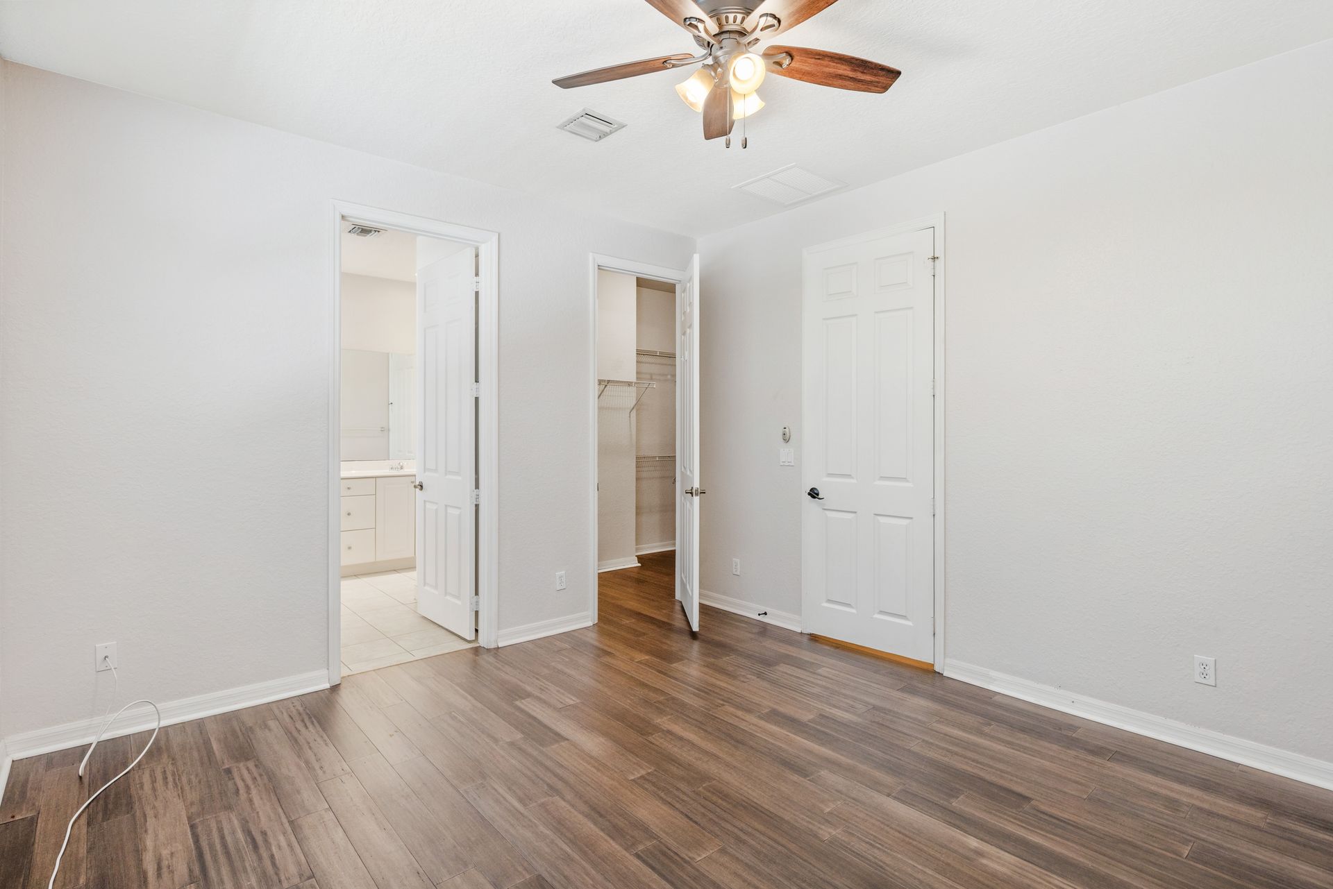 Empty bedroom with light grey walls, dark hardwood floors, a ceiling fan, and two open white doors leading to rooms.