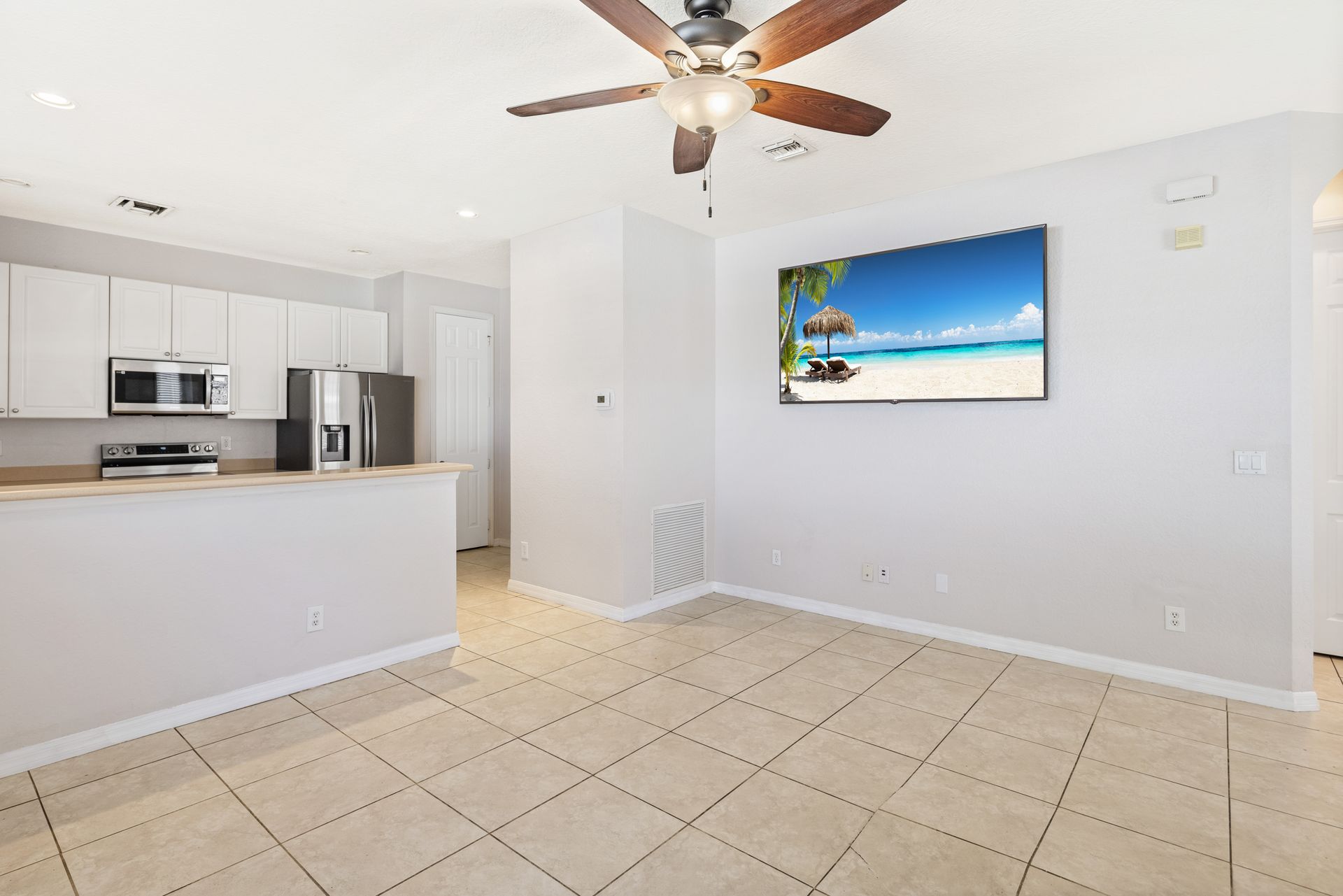 A brightly lit open-concept kitchen and living area with white cabinets, a ceiling fan, and a wall-mounted TV.