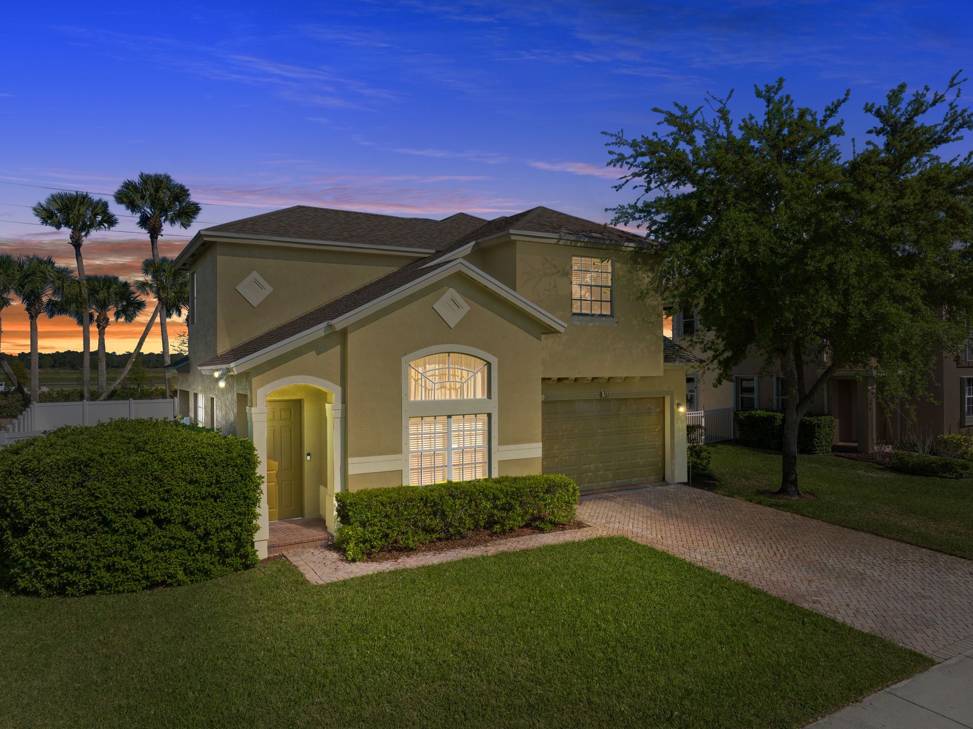 A two-story tan suburban house with a paved driveway and green lawn at sunset, featuring a small arched window.