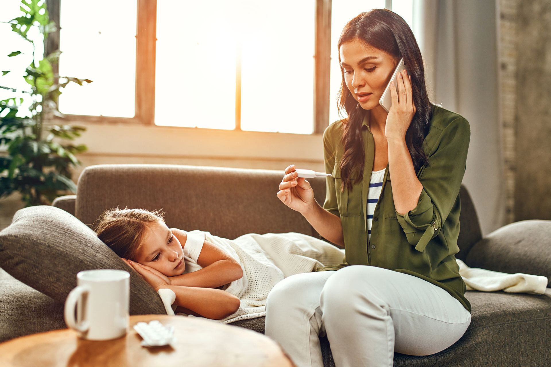 A woman is sitting on a couch with a sick child and talking on a cell phone.