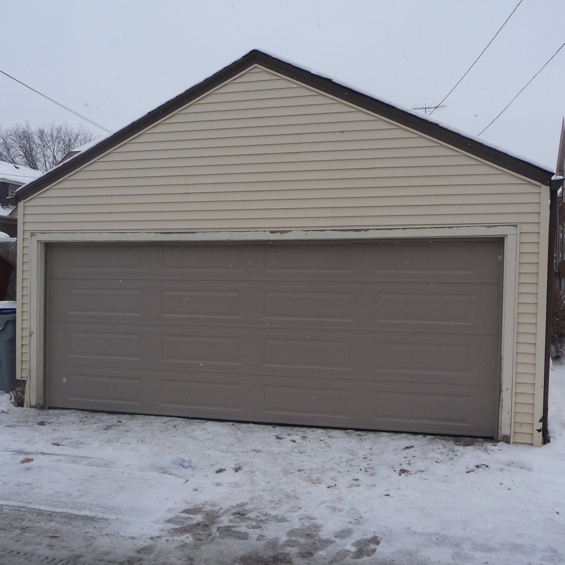 A detached garage with a closed gray door, beige siding, and a dark brown roof in a snowy setting.