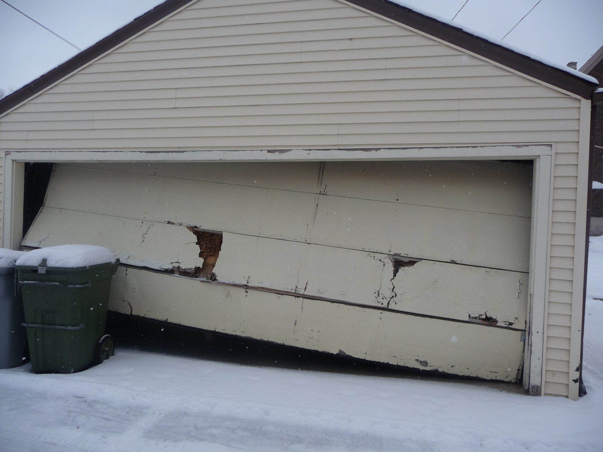 A damaged garage door, partially open, with visible structural damage and a broken section, set in a snowy environment.