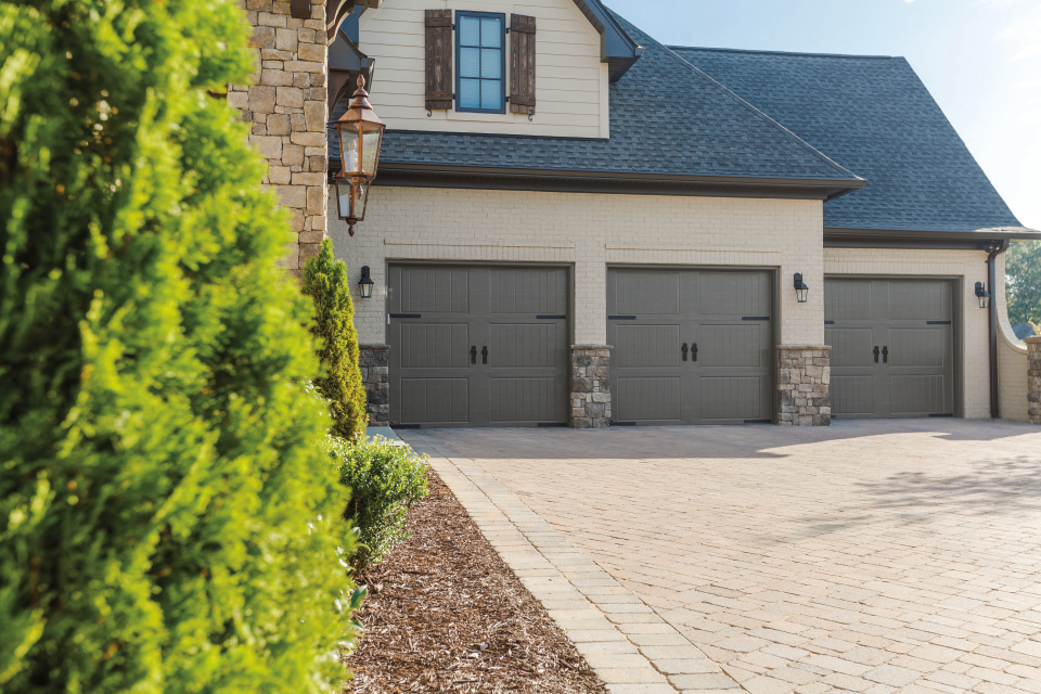 Exterior of a house with three tan garage doors, stone accents, and a brick driveway. Green bushes are in the foreground. 