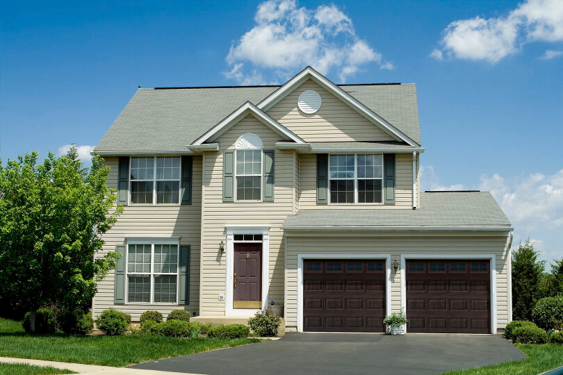 Two-story beige house with dark brown garage doors, green shutters, and a blue sky with clouds.