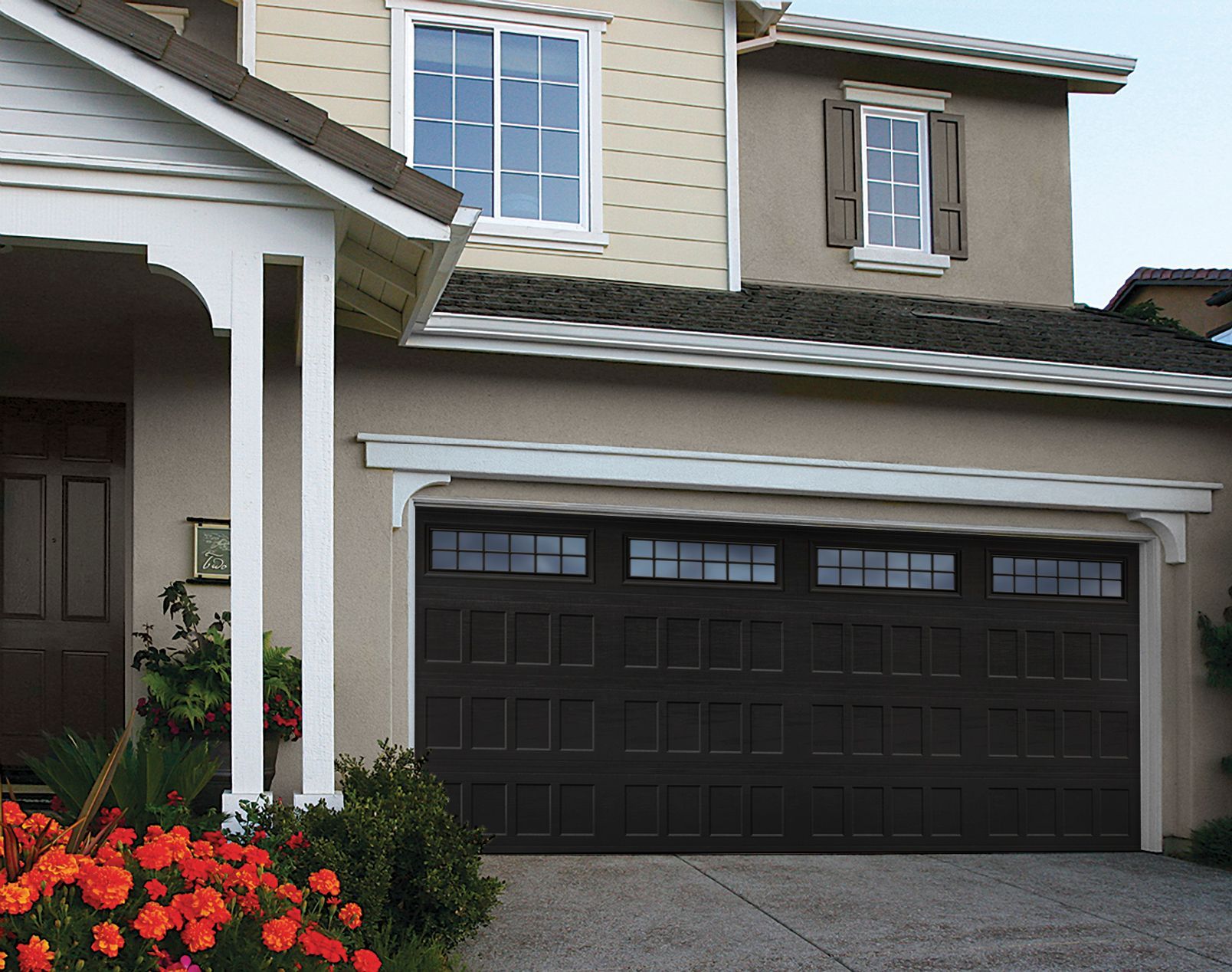 Brown garage door on a beige house with a white trim and windows, a flower bed in front.