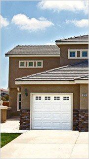 Two-story stucco house with a white garage door and a blue sky background. Brown roof, brick accents on front.