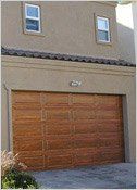 Tan stucco house with a brown wooden garage door. Two small white-framed windows are above.