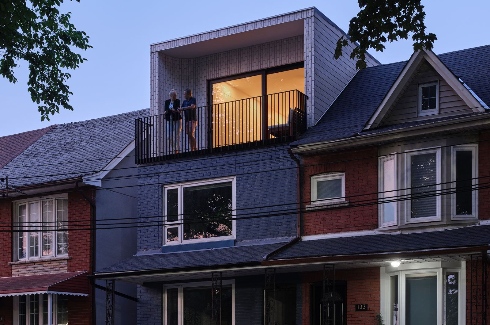 Modern home addition with balcony; two people overlooking street. Blue-gray siding, metal railings.