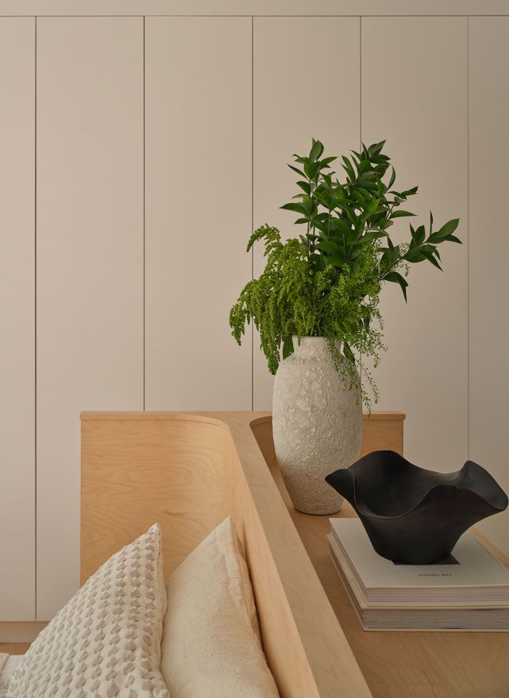 White vase with greenery, black bowl on books, and pillows on a light wood surface against white paneling.