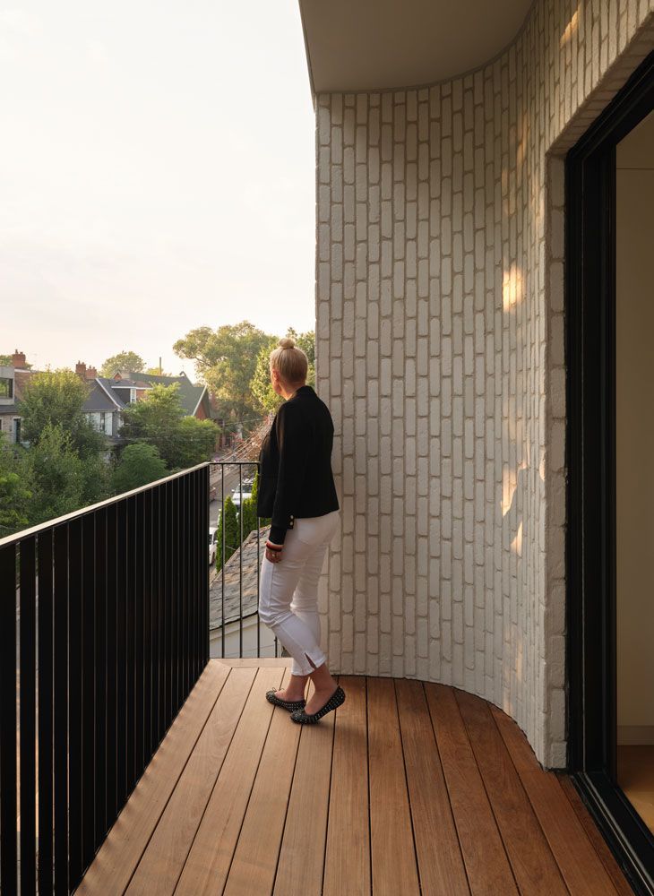 Woman on balcony looking at a tree-filled neighborhood; wood floor, black railing, white brick wall.