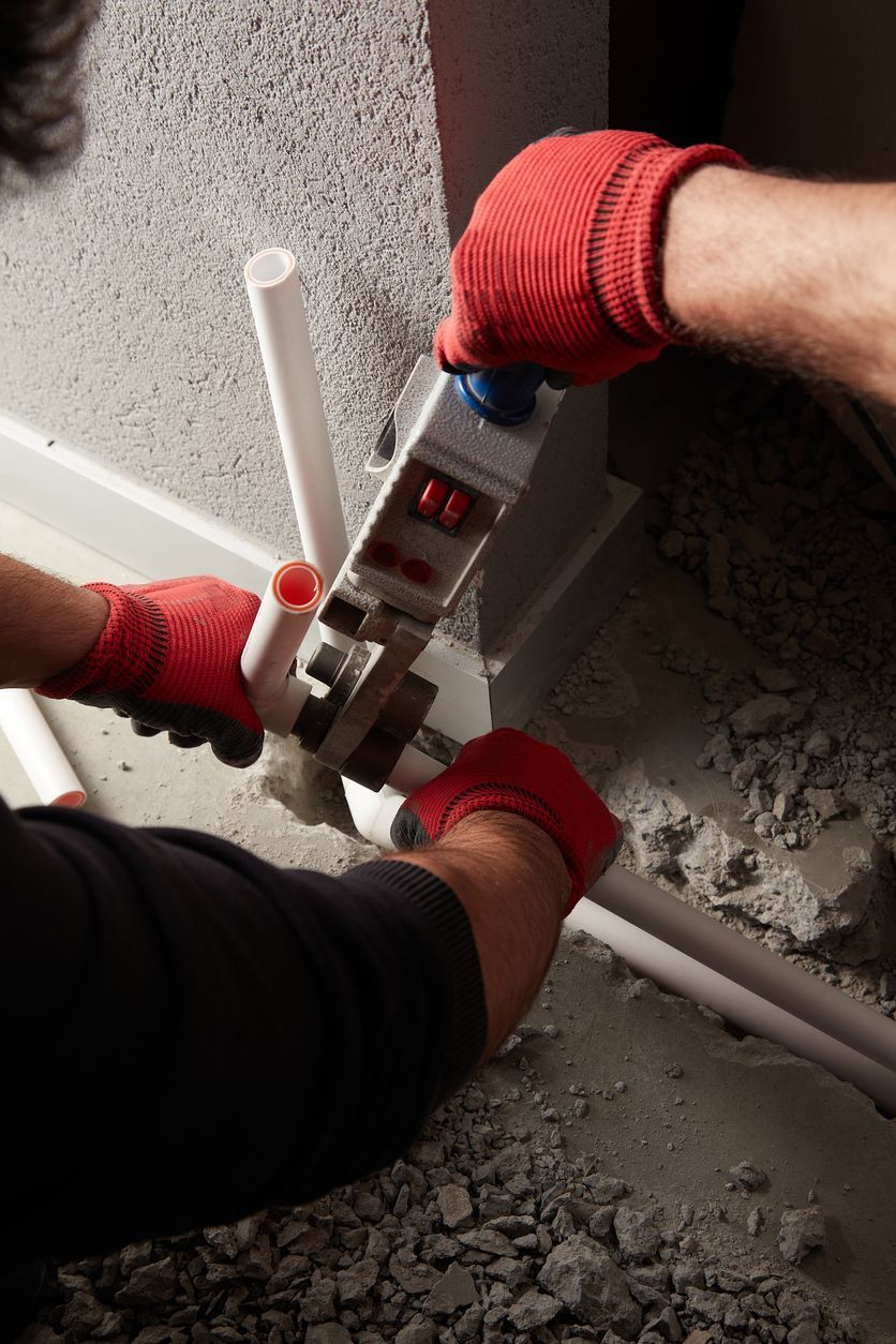 Gloved hands using a caulking gun to seal a white pipe along a rough wall.