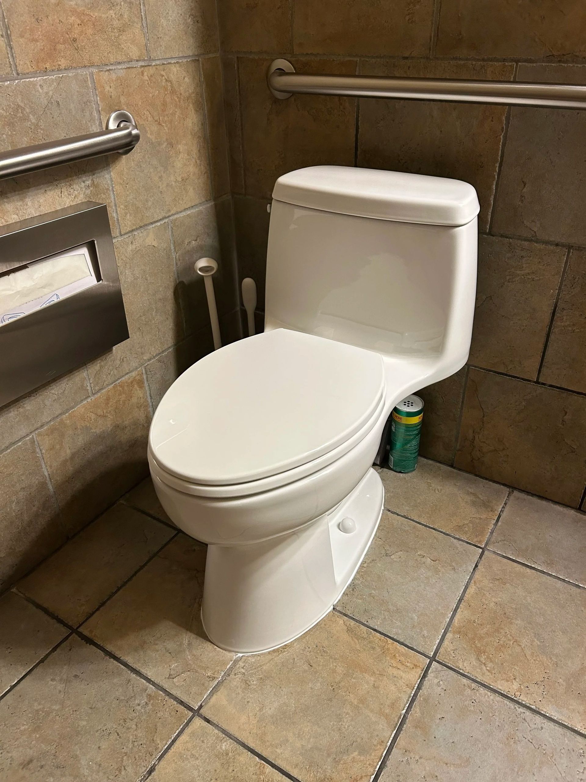 White toilet in a tiled public restroom with metal grab bars and a toilet paper dispenser.