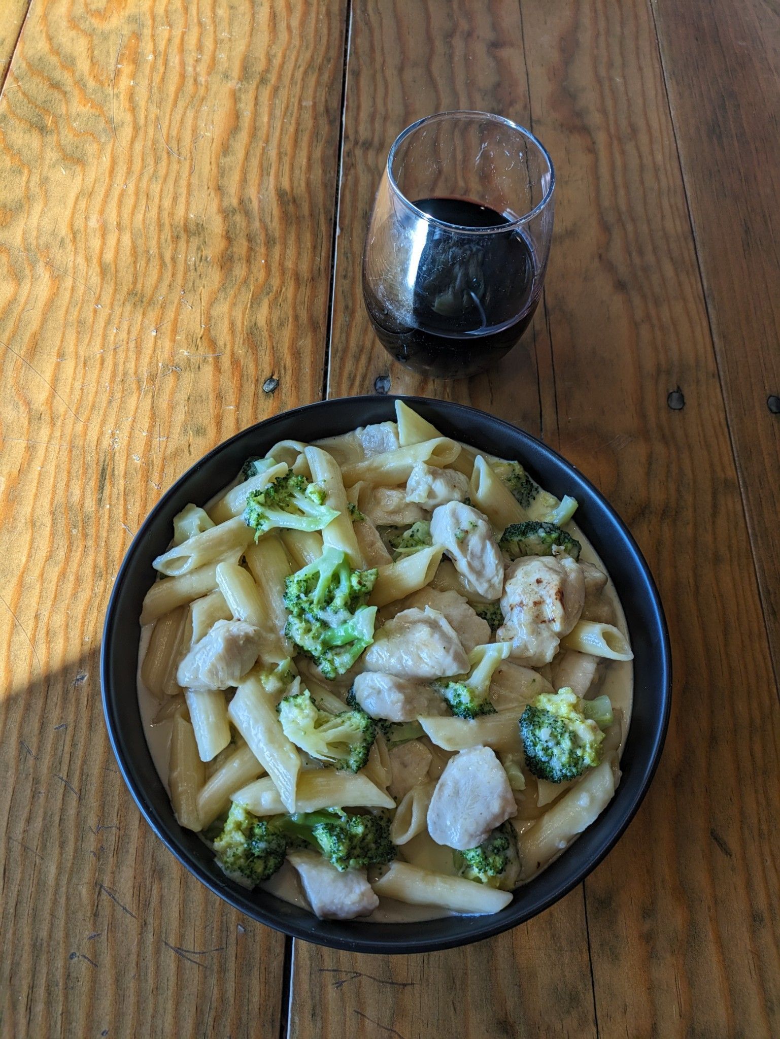 A bowl of pasta with broccoli and chicken next to a glass of wine on a wooden table.