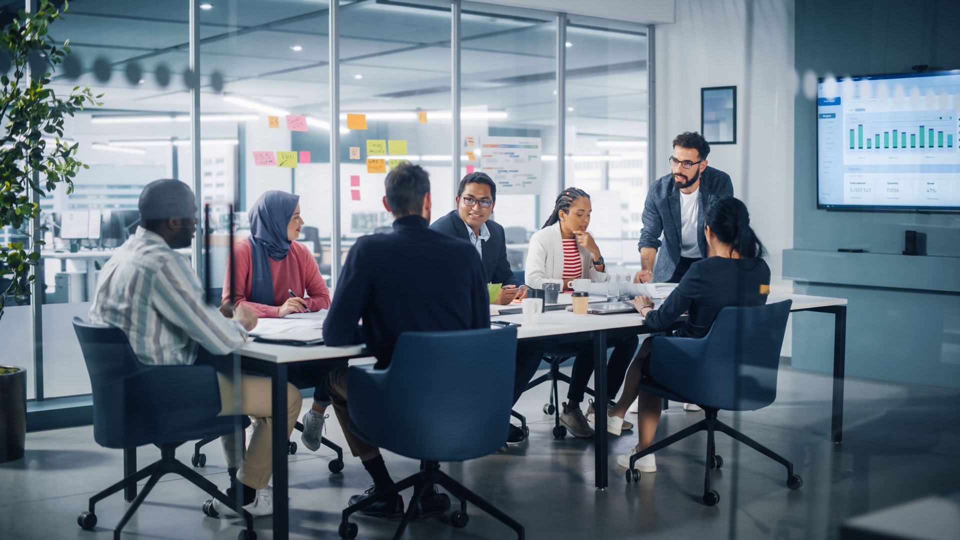 Business meeting in progress around a table in a modern office, with graphs on a display screen.