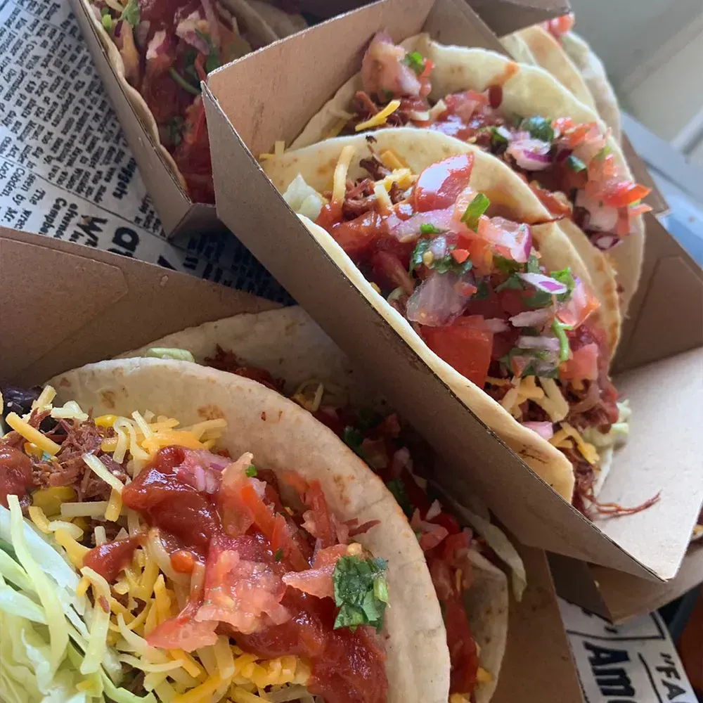 A bunch of tacos in a cardboard box with a newspaper in the background