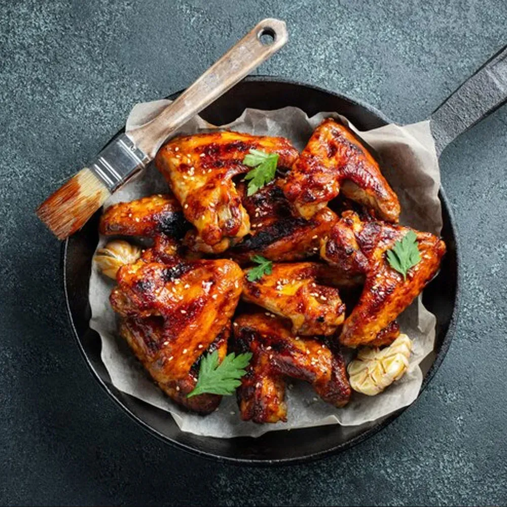A pan filled with chicken wings and a brush on a table.