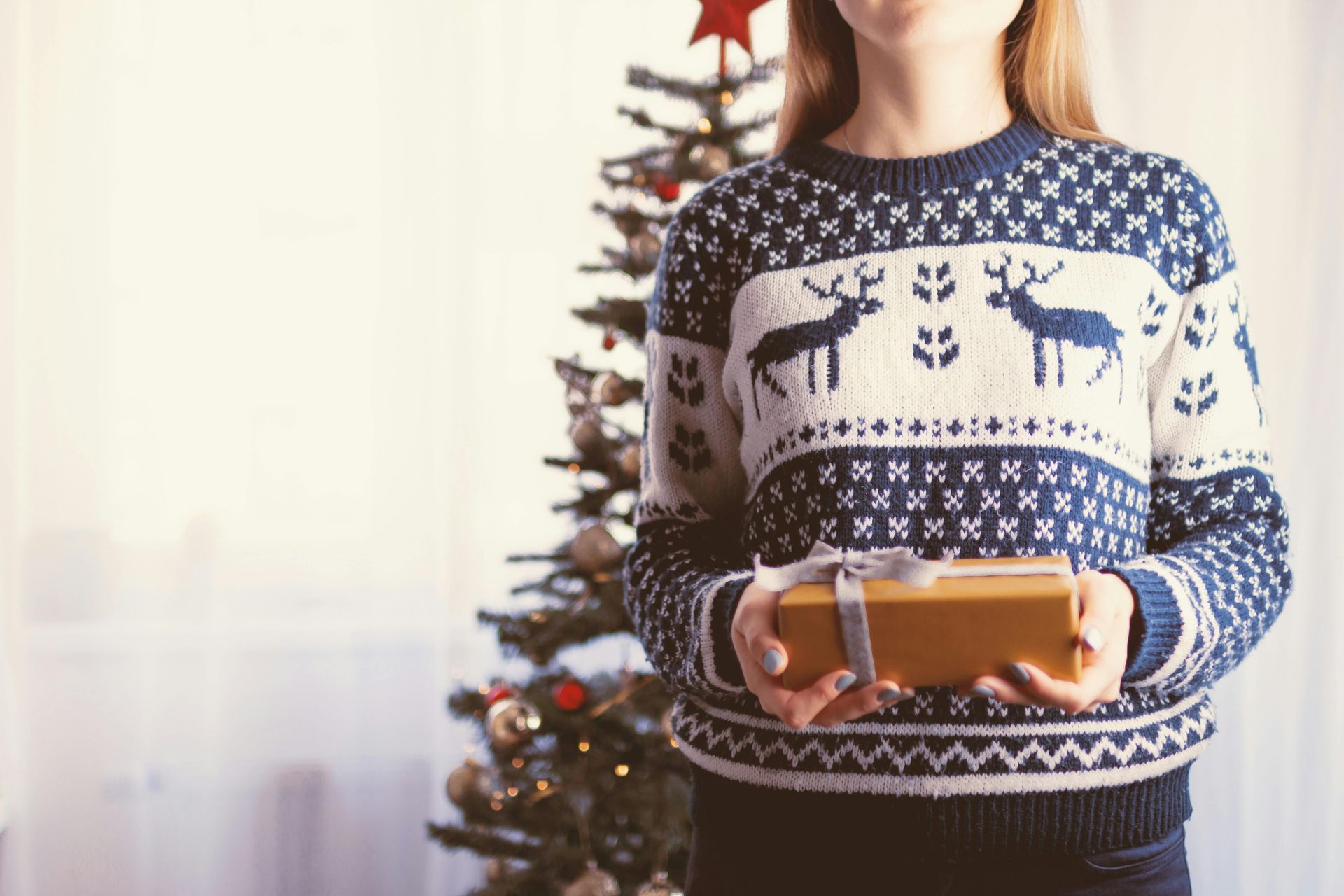 A woman in a sweater is holding a christmas present in front of a christmas tree.