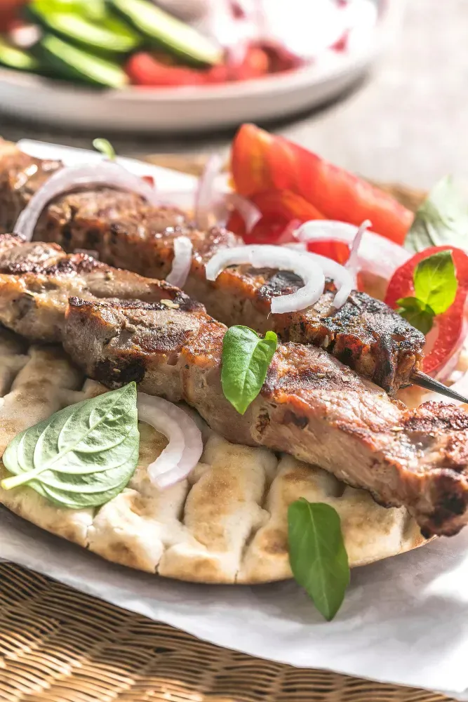 A close up of a plate of food with Lamb Souvlaki meat and vegetables on a table.