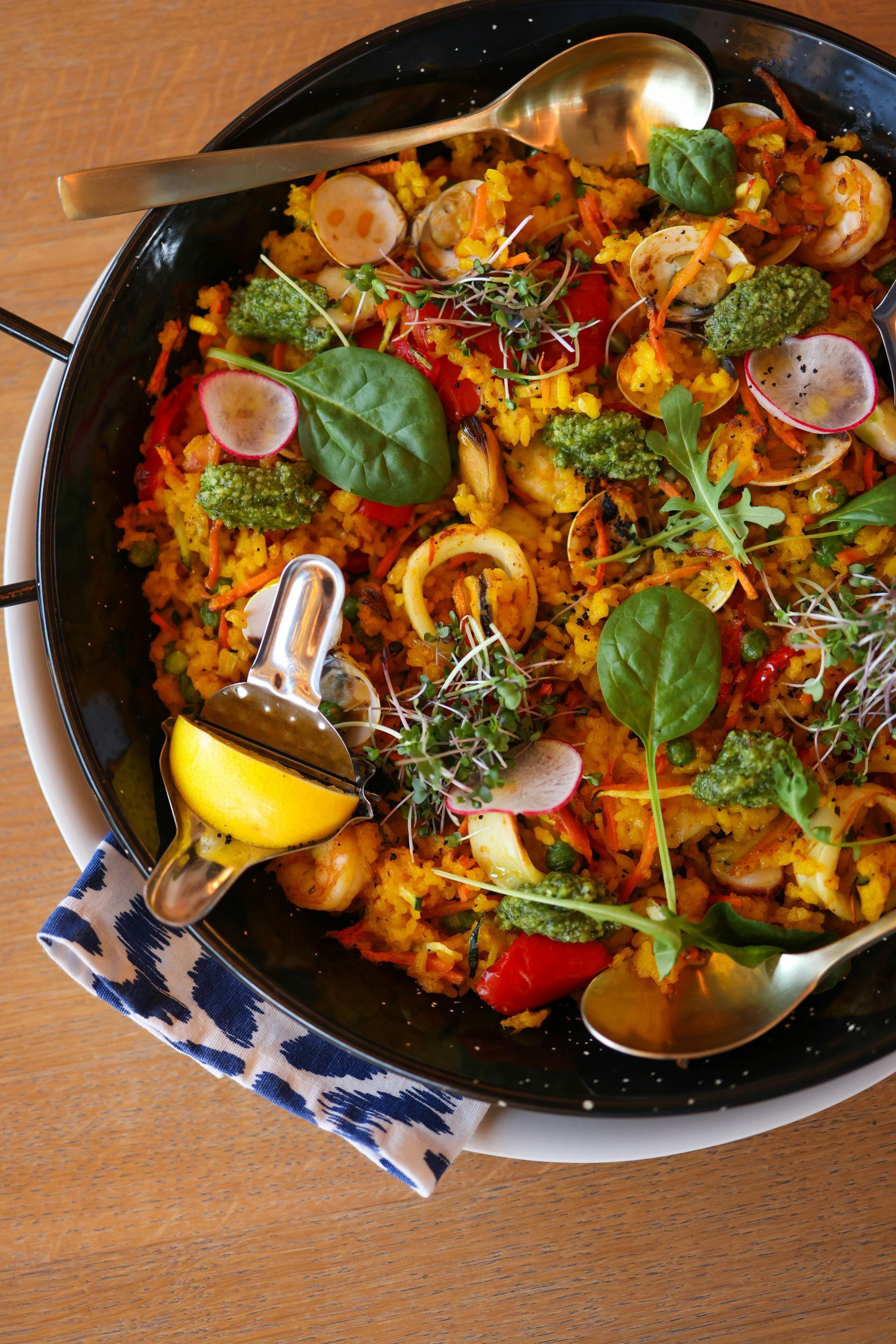 A pan of Paella food with a spoon and a lemon on a table.