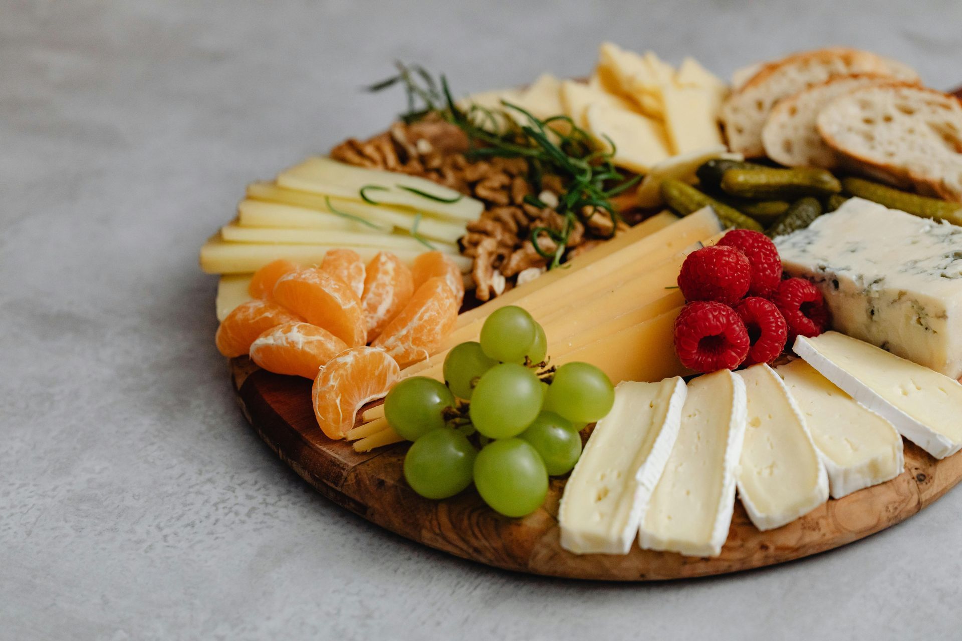 A wooden cutting board topped with cheese , grapes , raspberries , oranges and nuts.