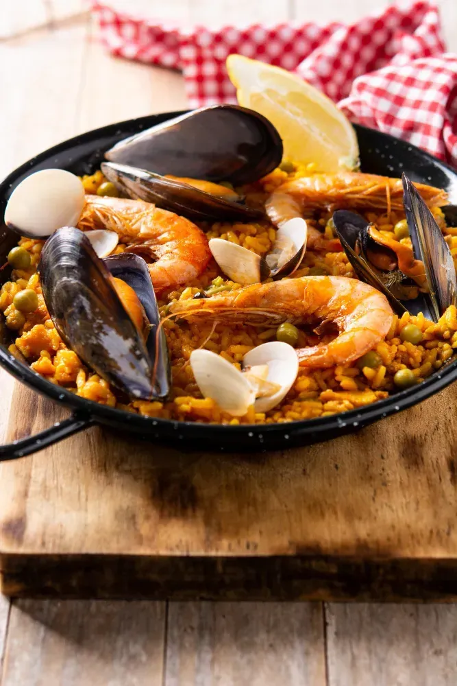A pan of paella with shrimp , mussels and rice on a wooden cutting board.