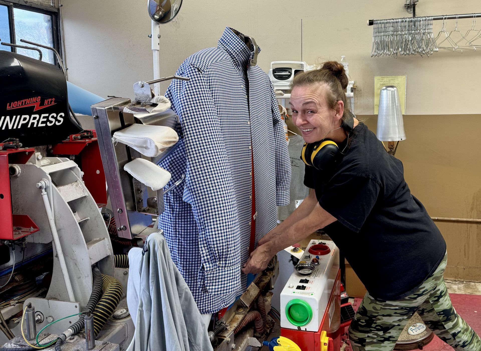 A person in a black shirt works at a professional Unipress clothing machine, adjusting a blue and white checkered shirt.