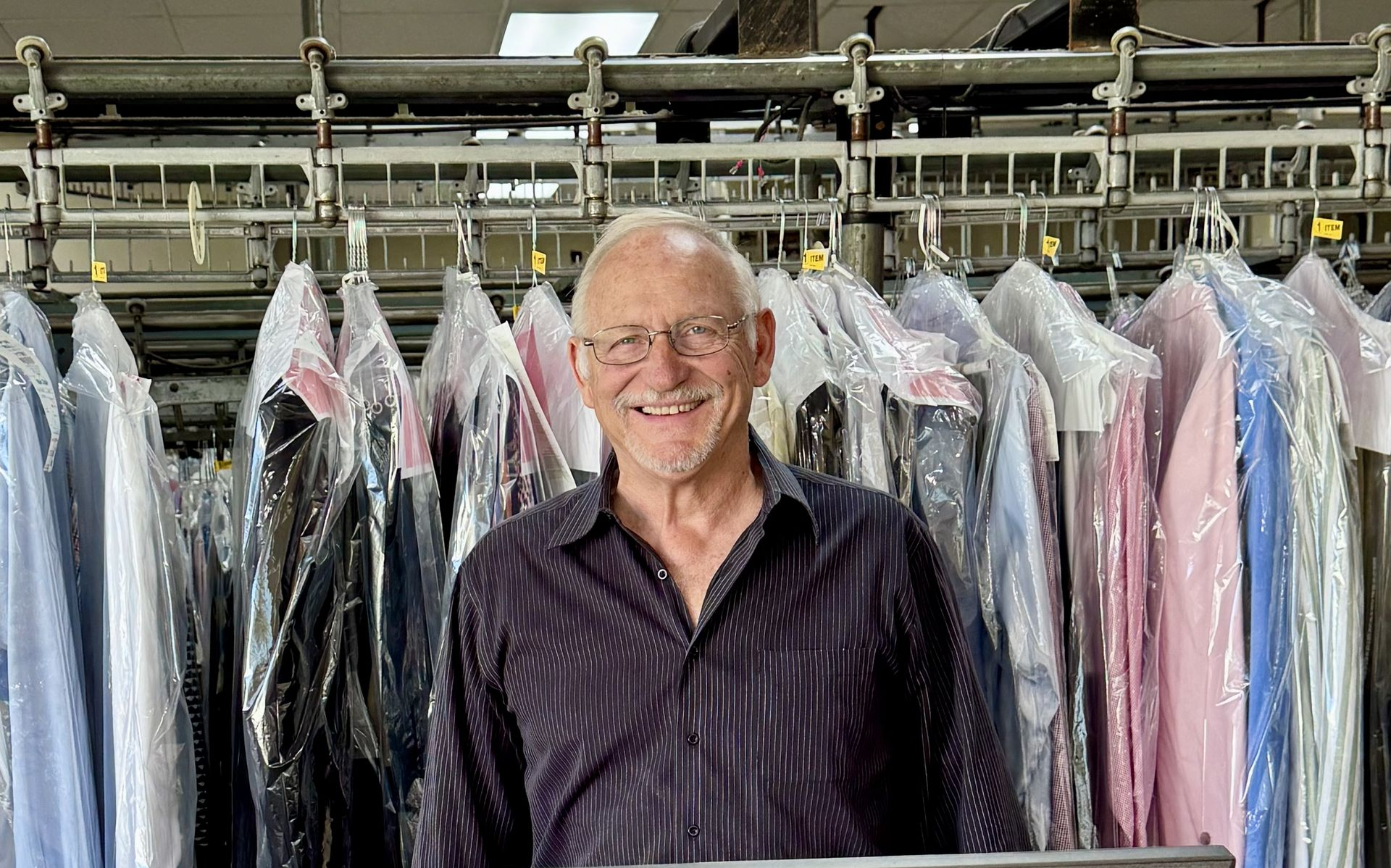 Smiling person in a dark shirt standing in front of rows of clothes wrapped in plastic on a conveyor rack.