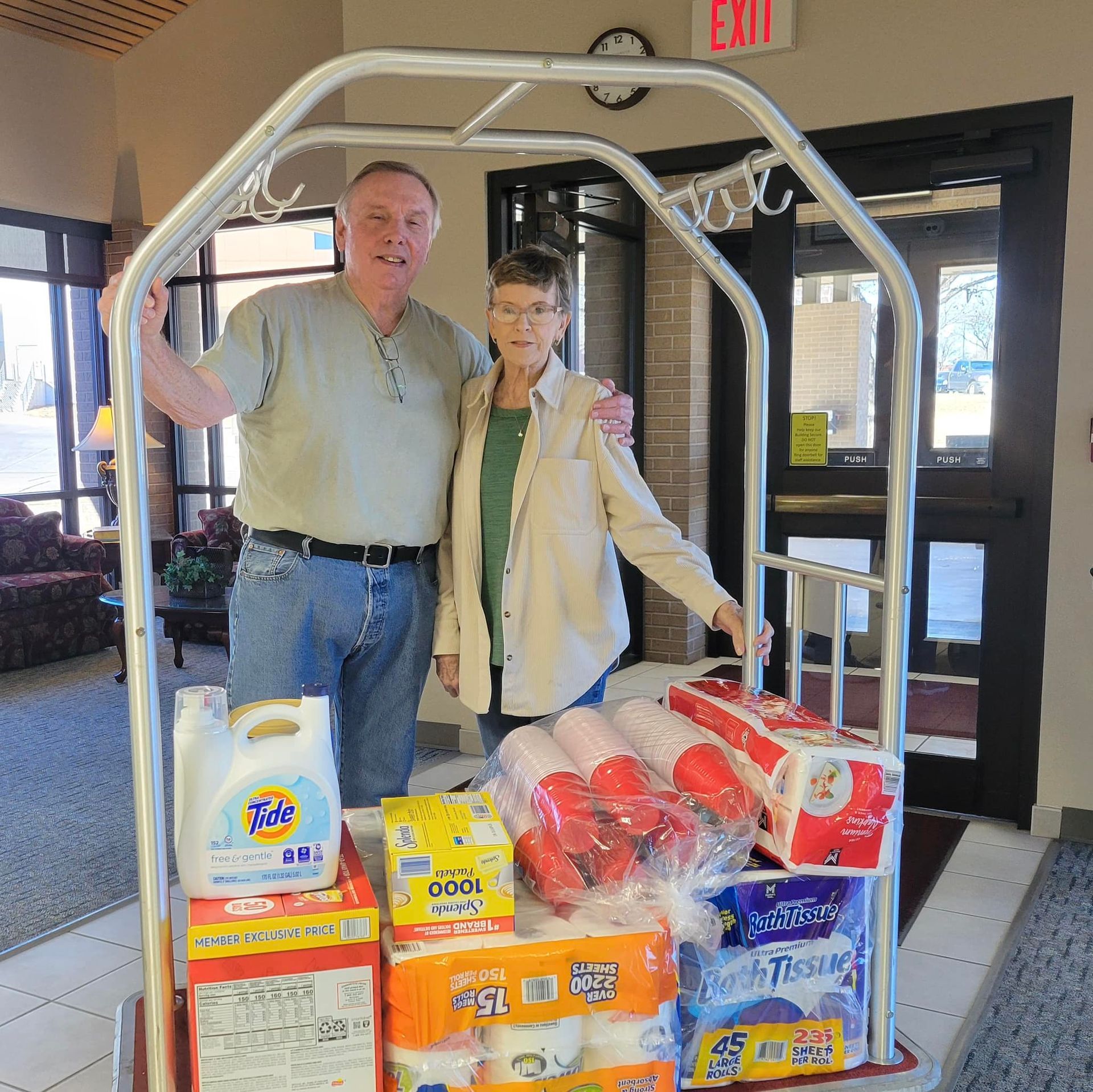 Man and woman stand by cart loaded with supplies. Inside a building with an exit sign.
