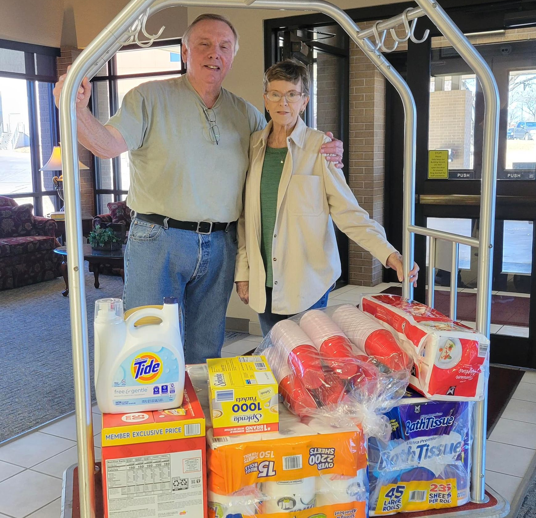Couple with supplies in hotel. Man with arm around woman, standing by a cart of donations like detergent and paper towels.