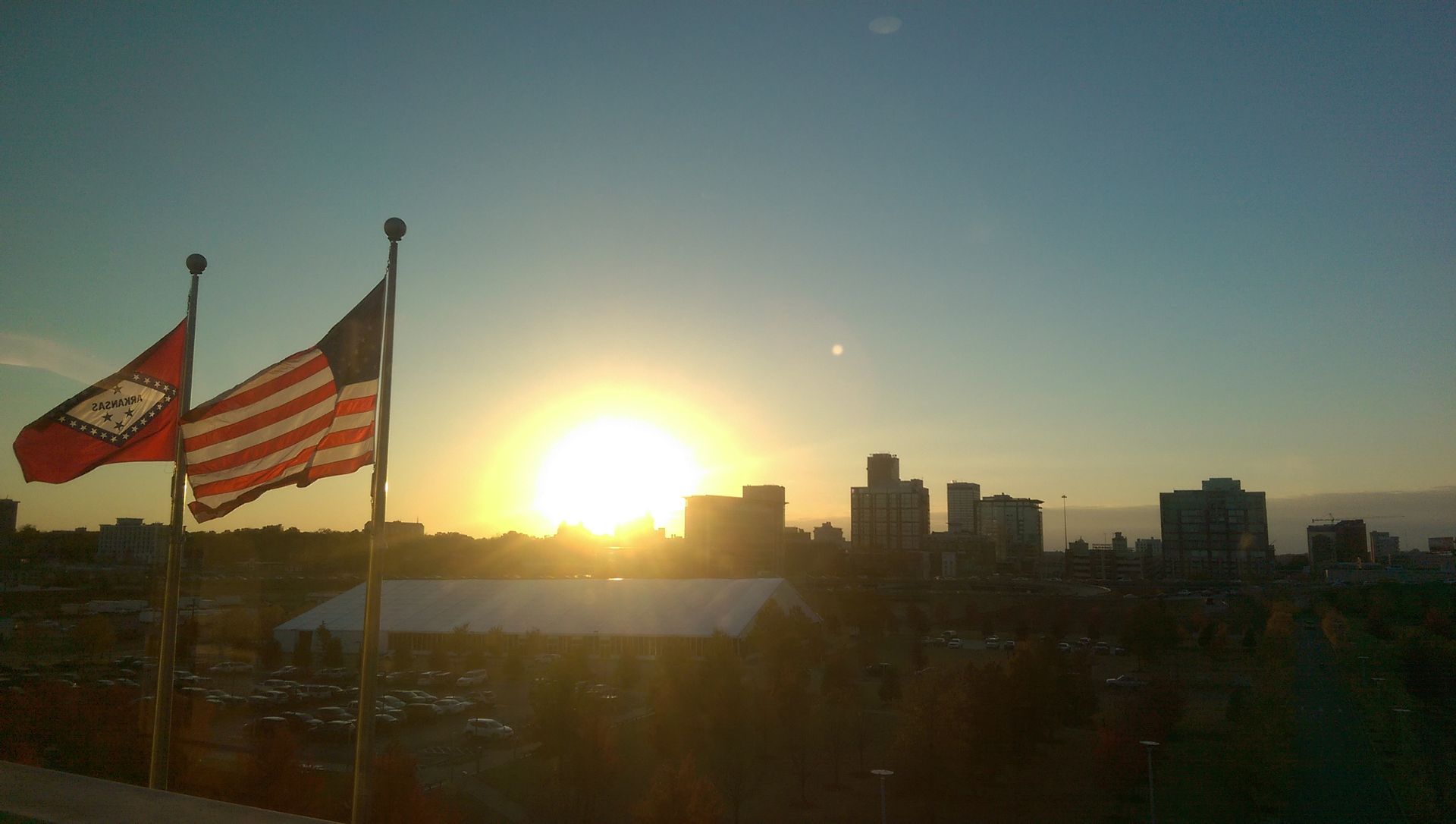 Two flags are flying in front of a sunset over a city