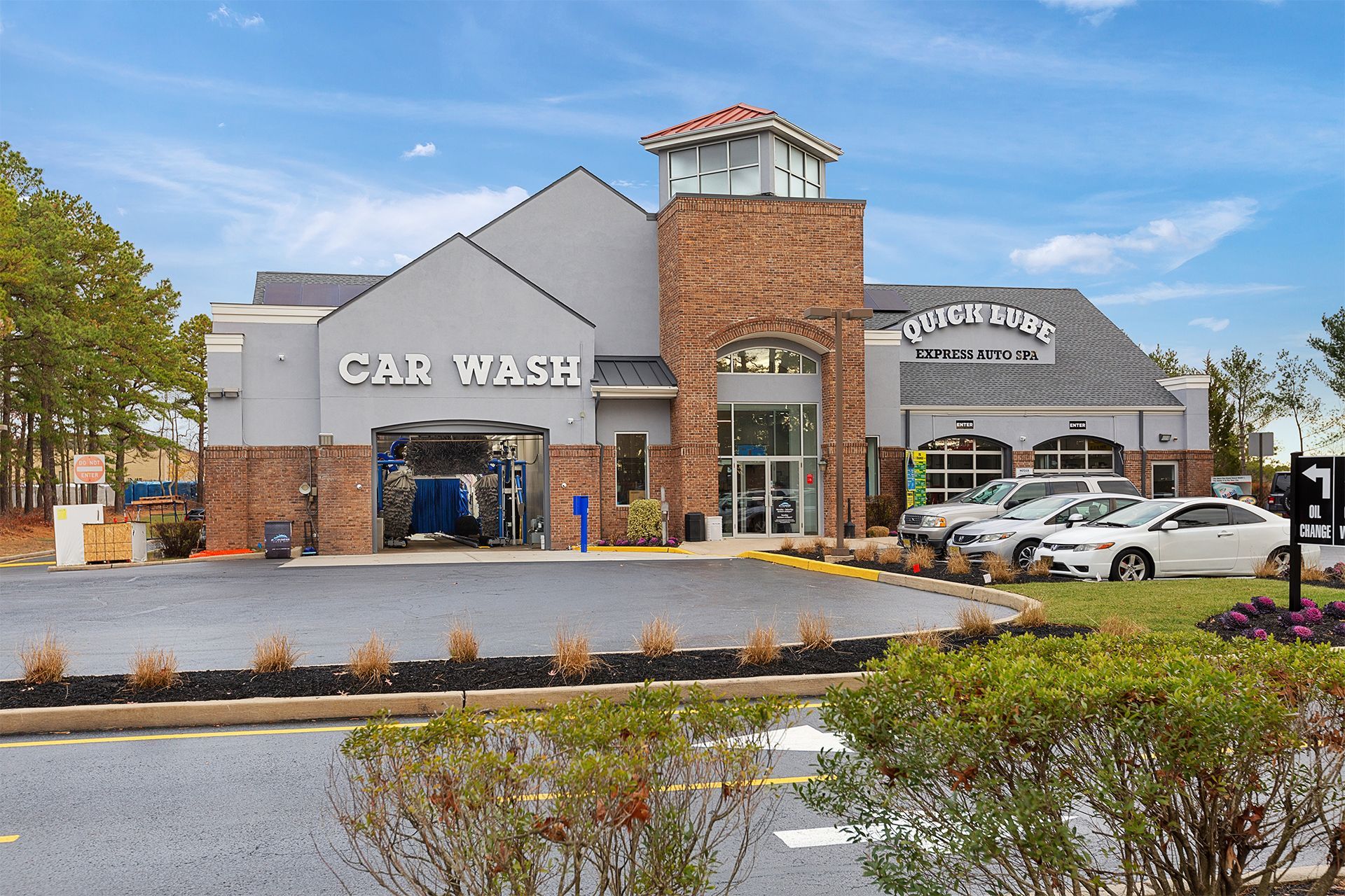 Exterior of a car wash facility with a brick and gray facade. Cars parked in front, clear blue sky.