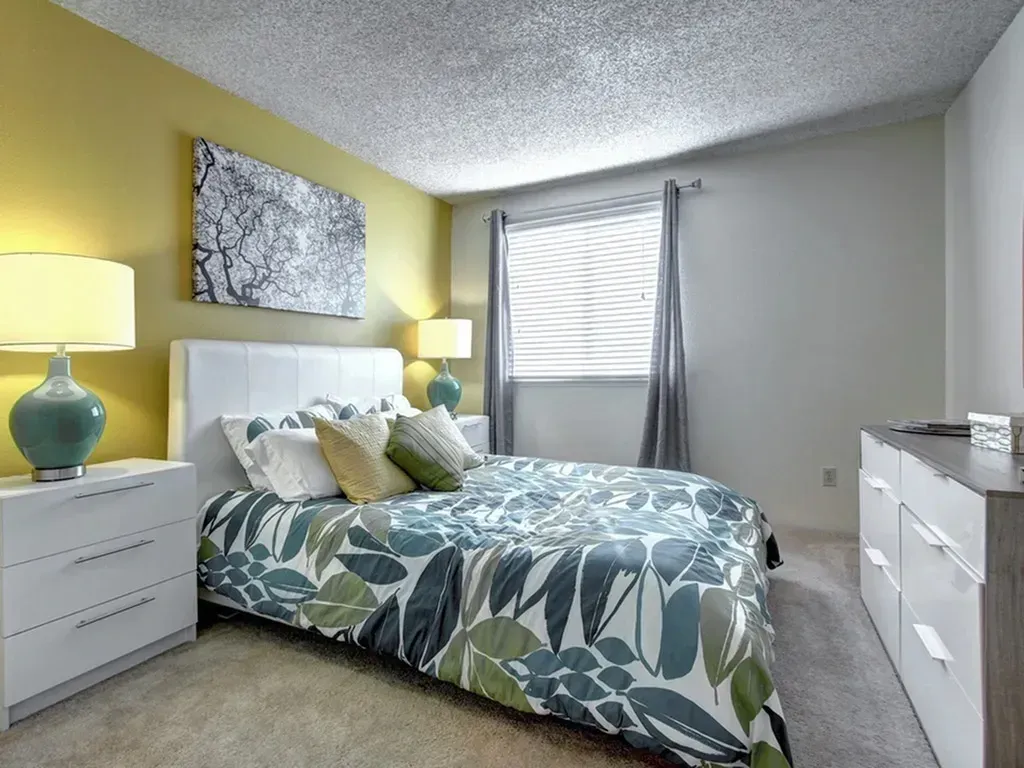 Bright bedroom with yellow and white walls, floral bedspread, two lamps, and a window with blinds.