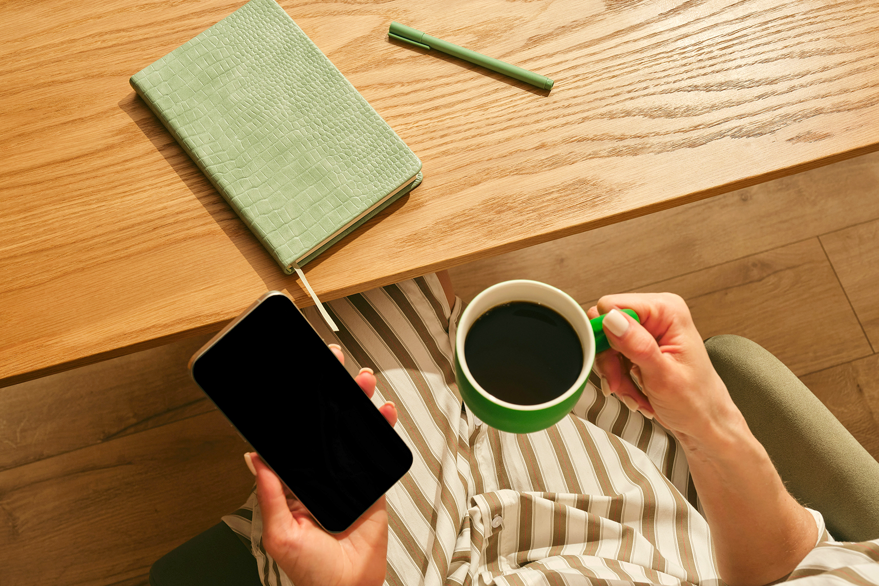 Person holding a phone and mug at a desk with a notebook and pencil on a wooden floor.