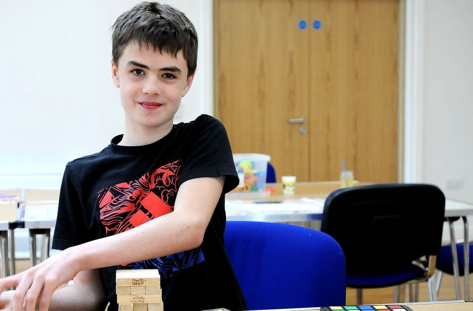 Teen smiles, leaning on wooden blocks. Black t-shirt with red design. Indoor setting, light wood door.