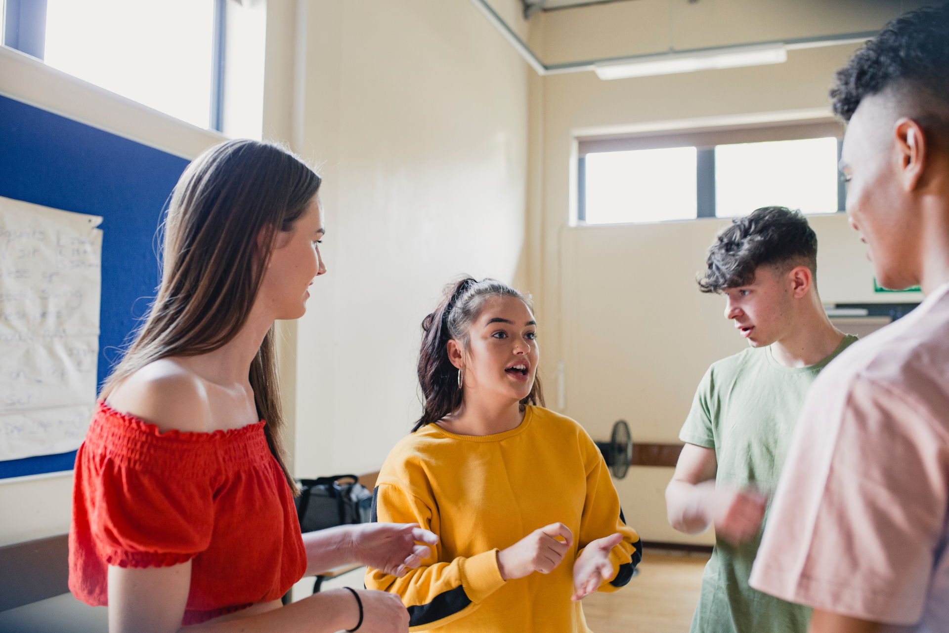 Four young people in a room, talking and gesturing. One in a red top, another in yellow.