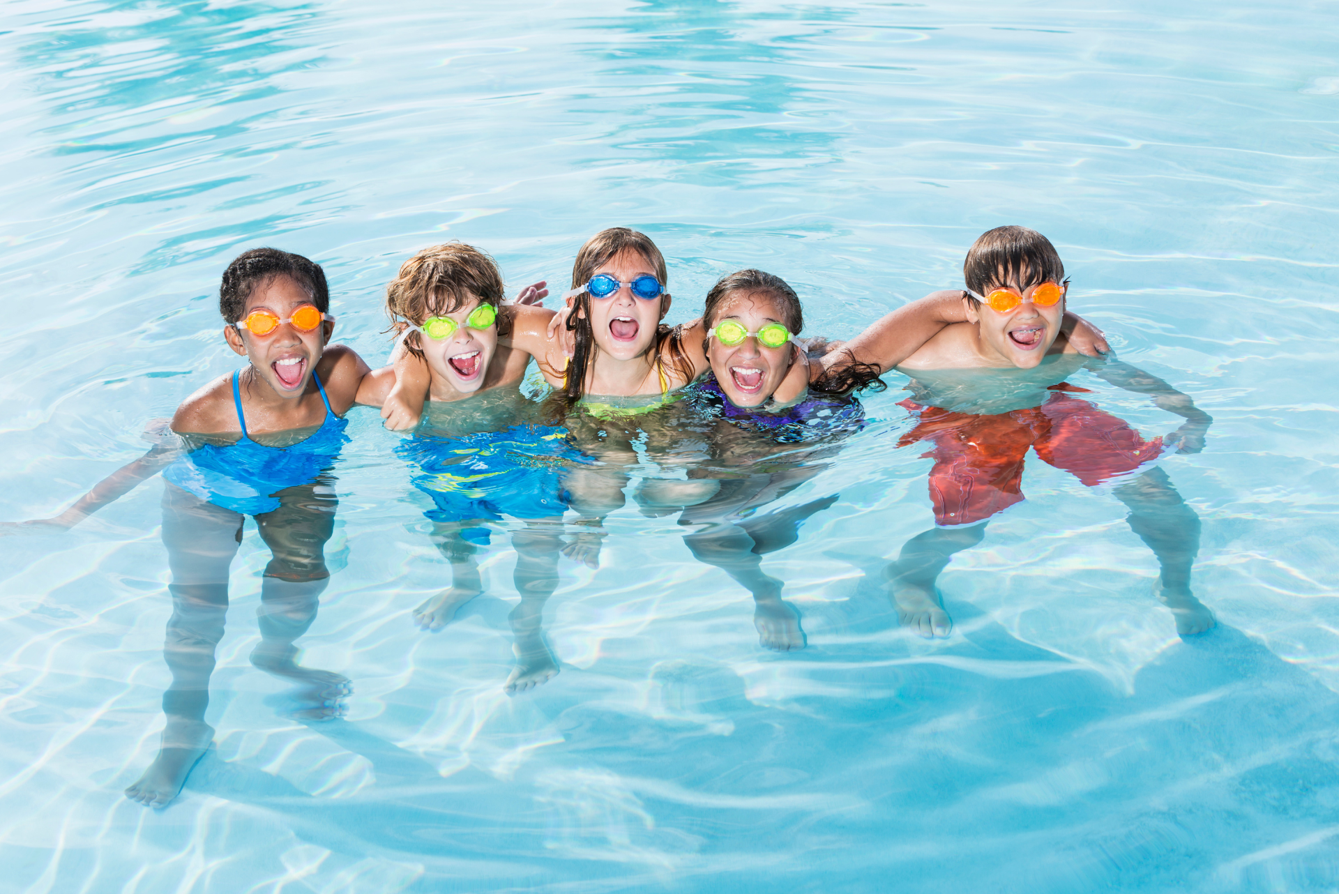 Five children wearing goggles, smiling, and posing in a pool.