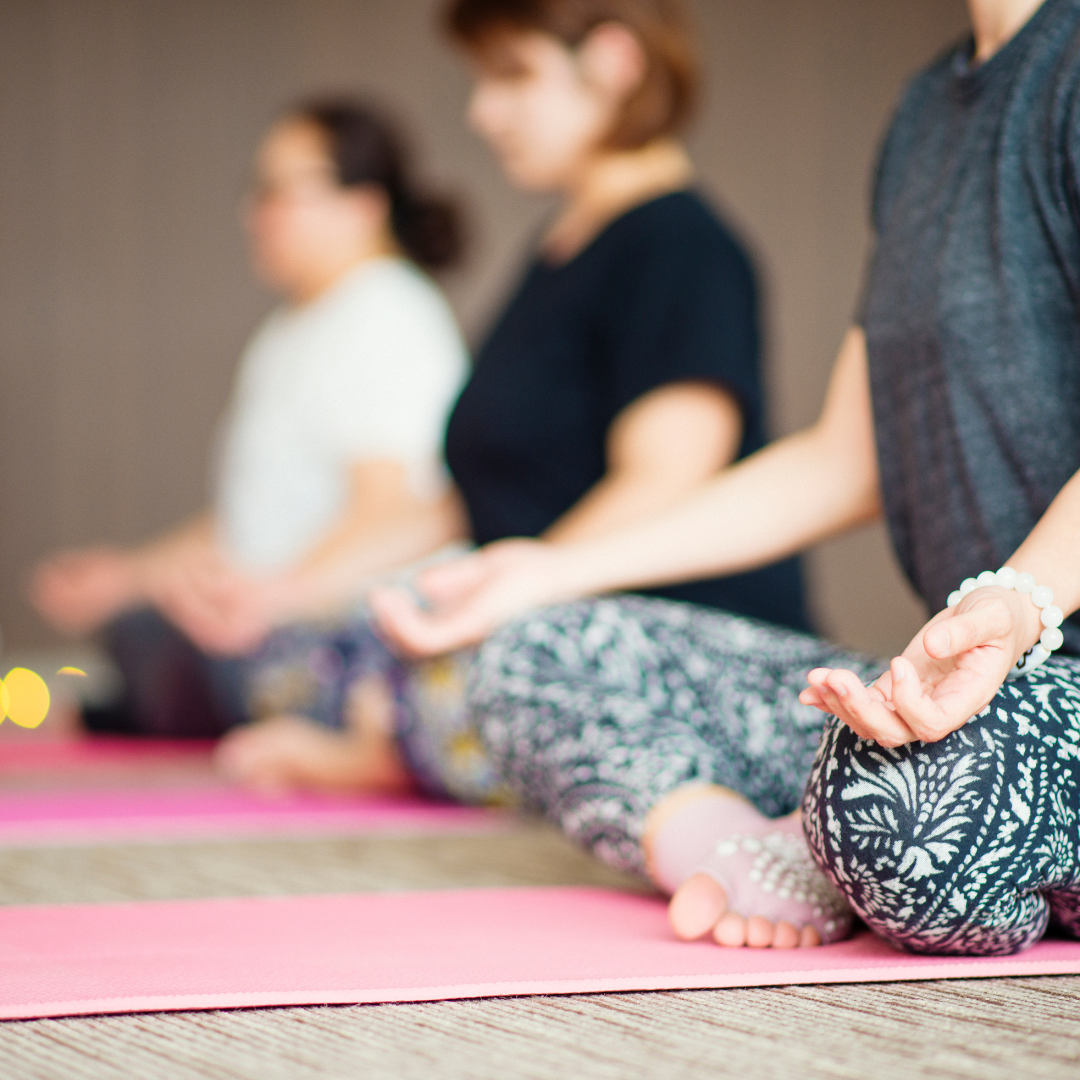 Three people in a yoga class seated in lotus position on pink mats, eyes closed, hands in their laps.
