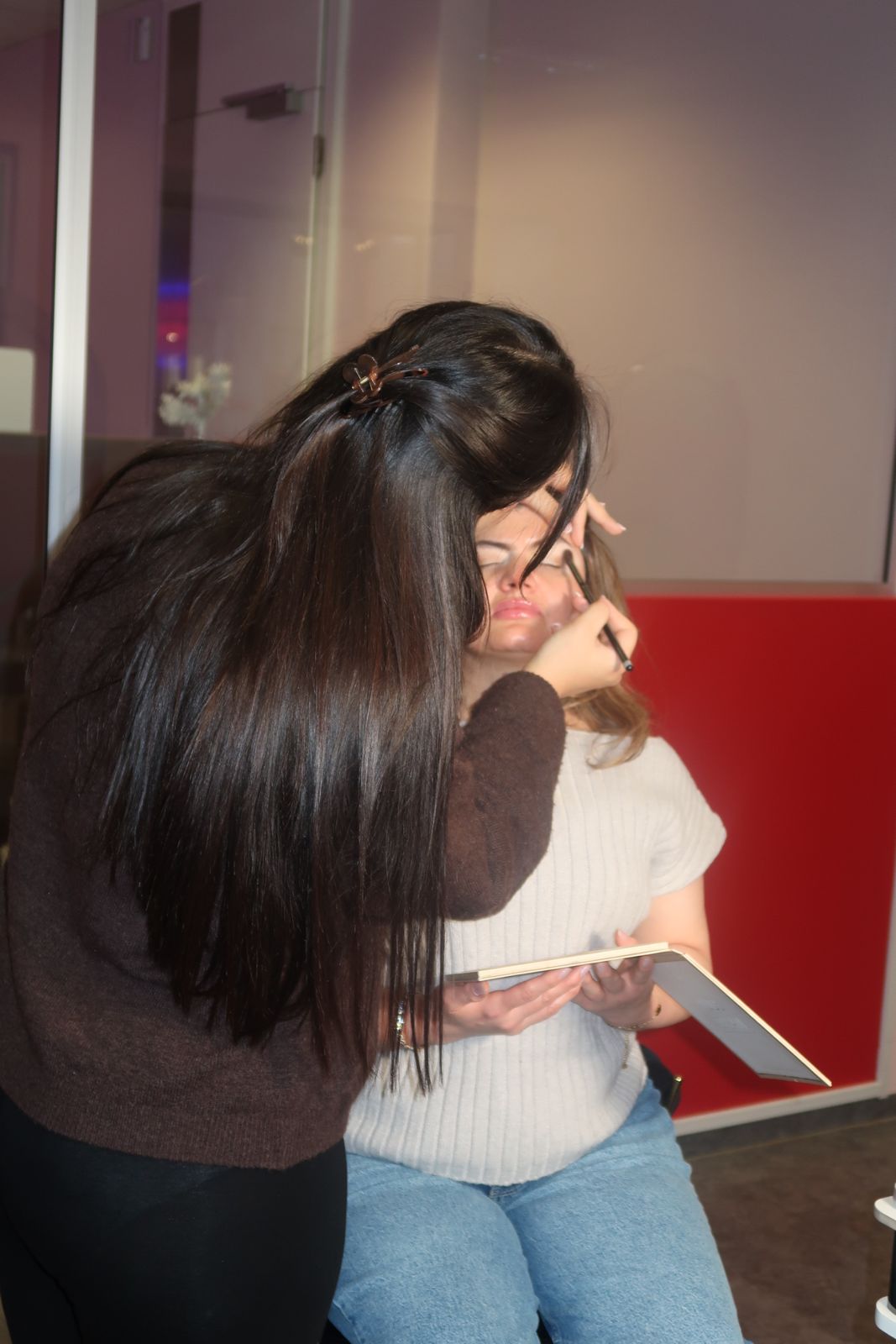 Eine Frau schminkt einer anderen Frau das Gesicht. Beide befinden sich in einem Gebäude, eine hält eine Palette in der Hand.