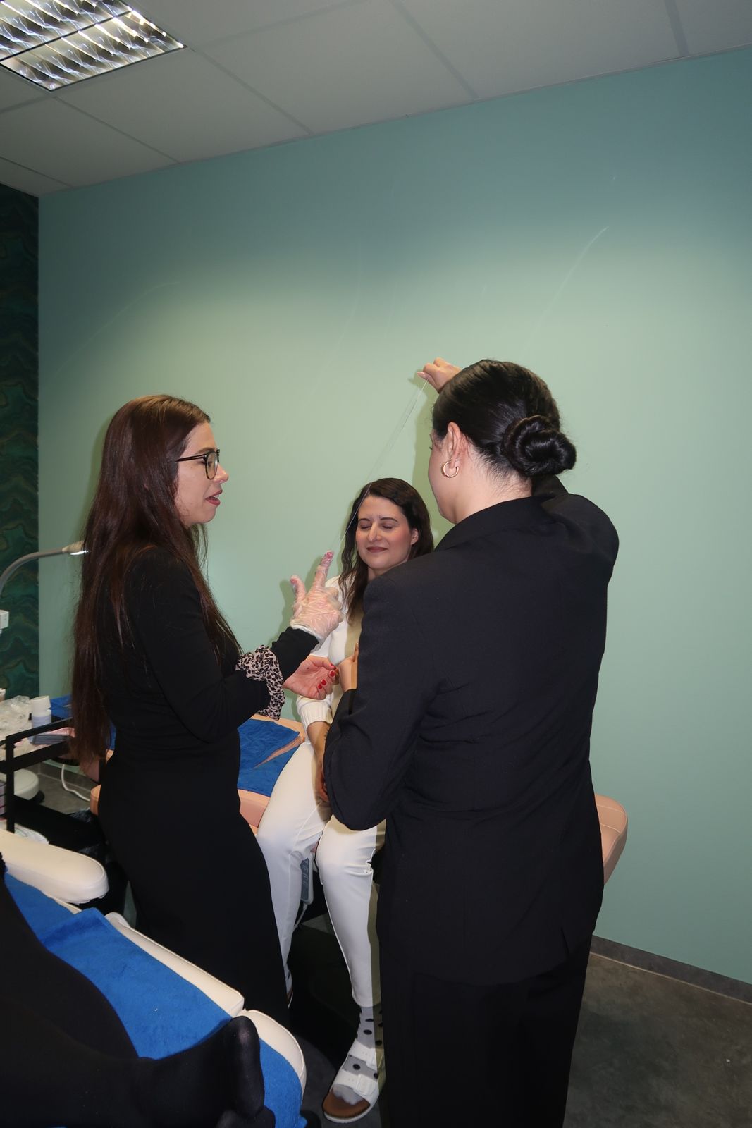 Drei Personen in einem Raum; zwei stehen, eine sitzt. Eine Frau hält ein durchsichtiges Glas; die anderen beiden stehen ihr gegenüber.