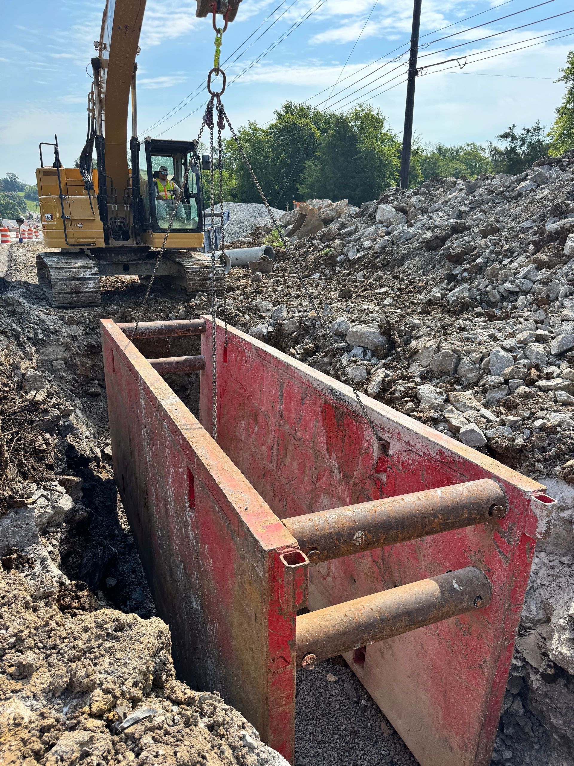 Construction site: excavator lifting a red trench box into a trench. Dirt and rocks surround the trench.