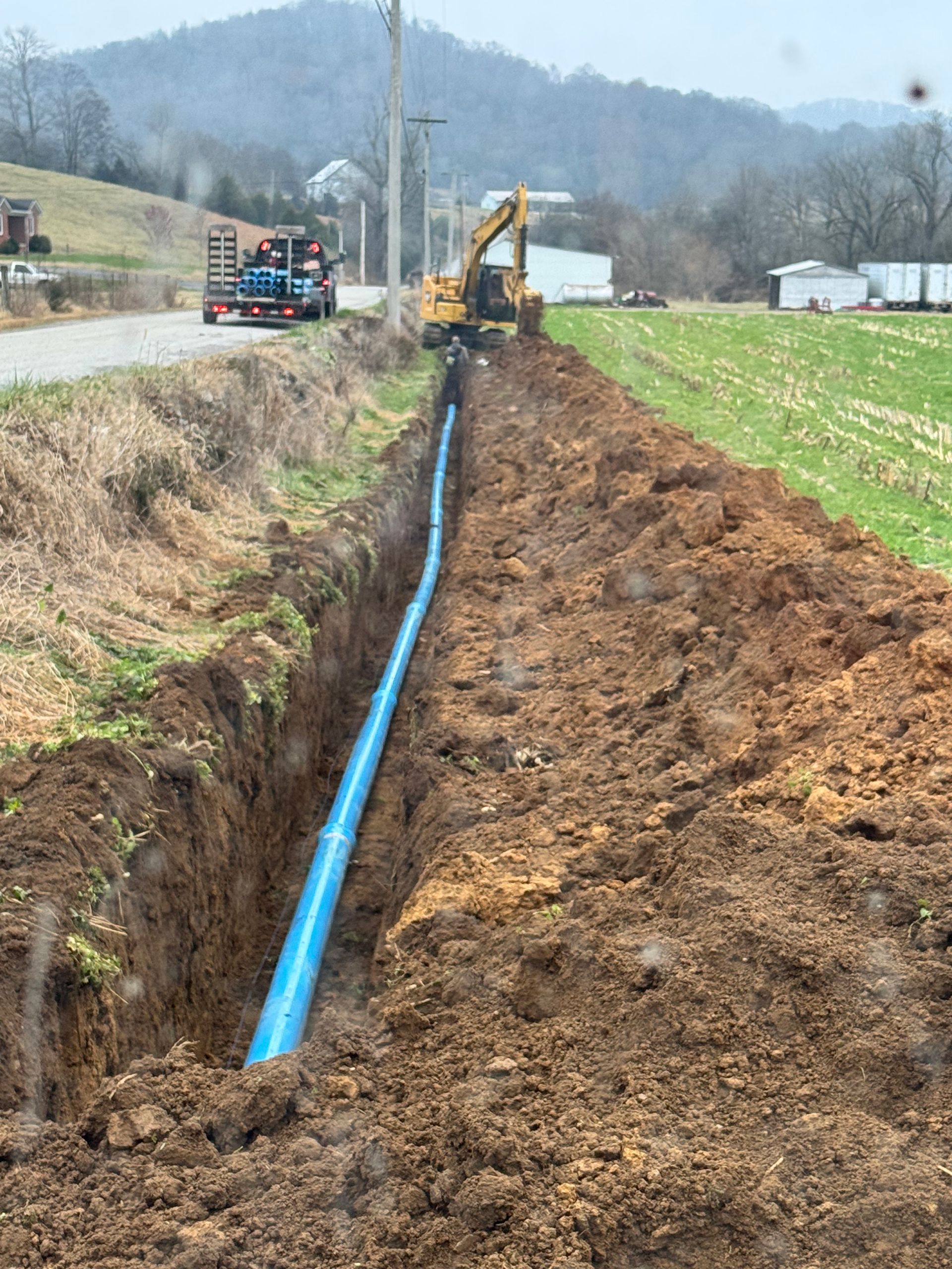 A blue water pipe being installed in a trench alongside a rural road by an excavator.