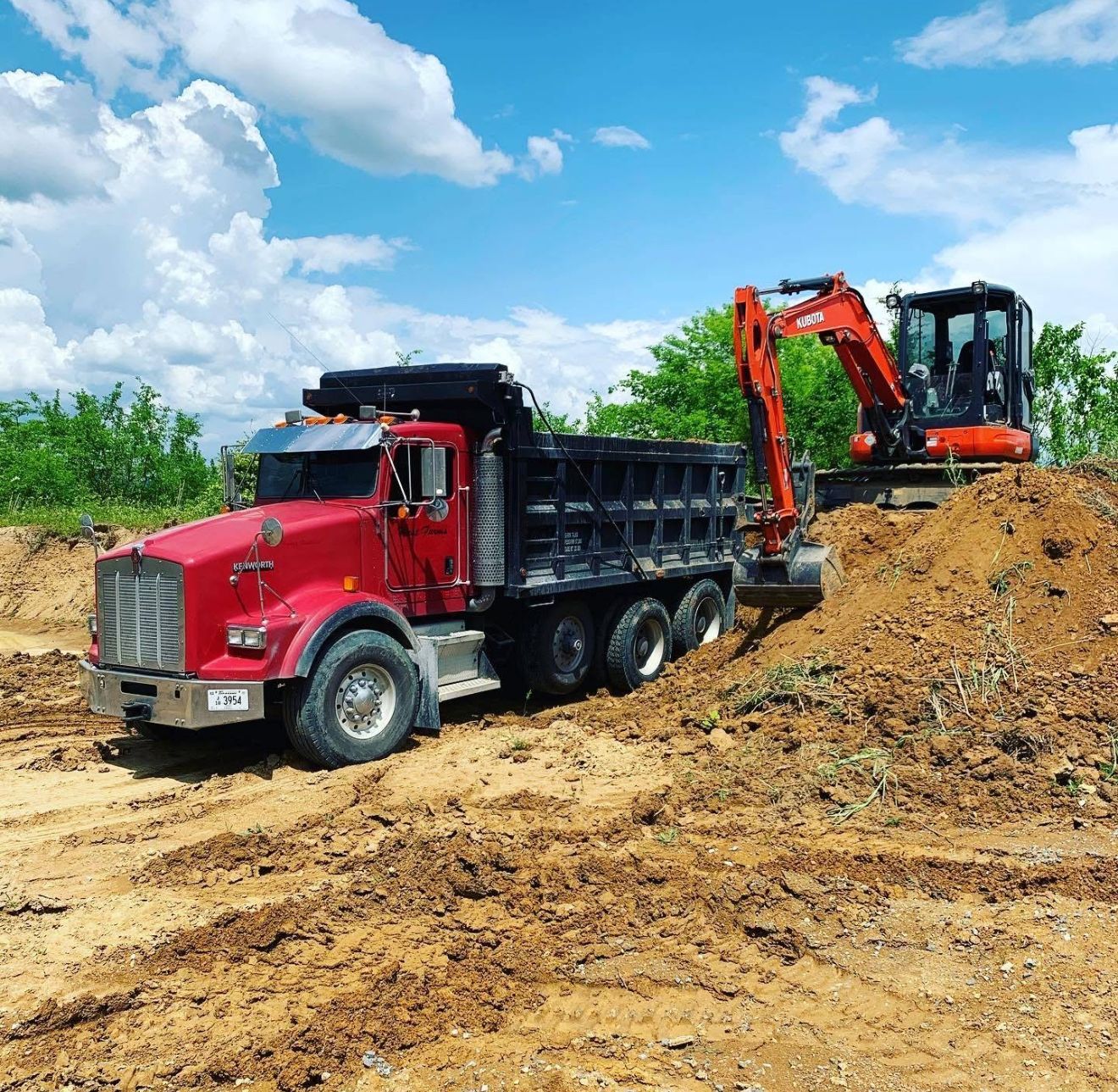A red dump truck is driving down a dirt road next to an excavator.