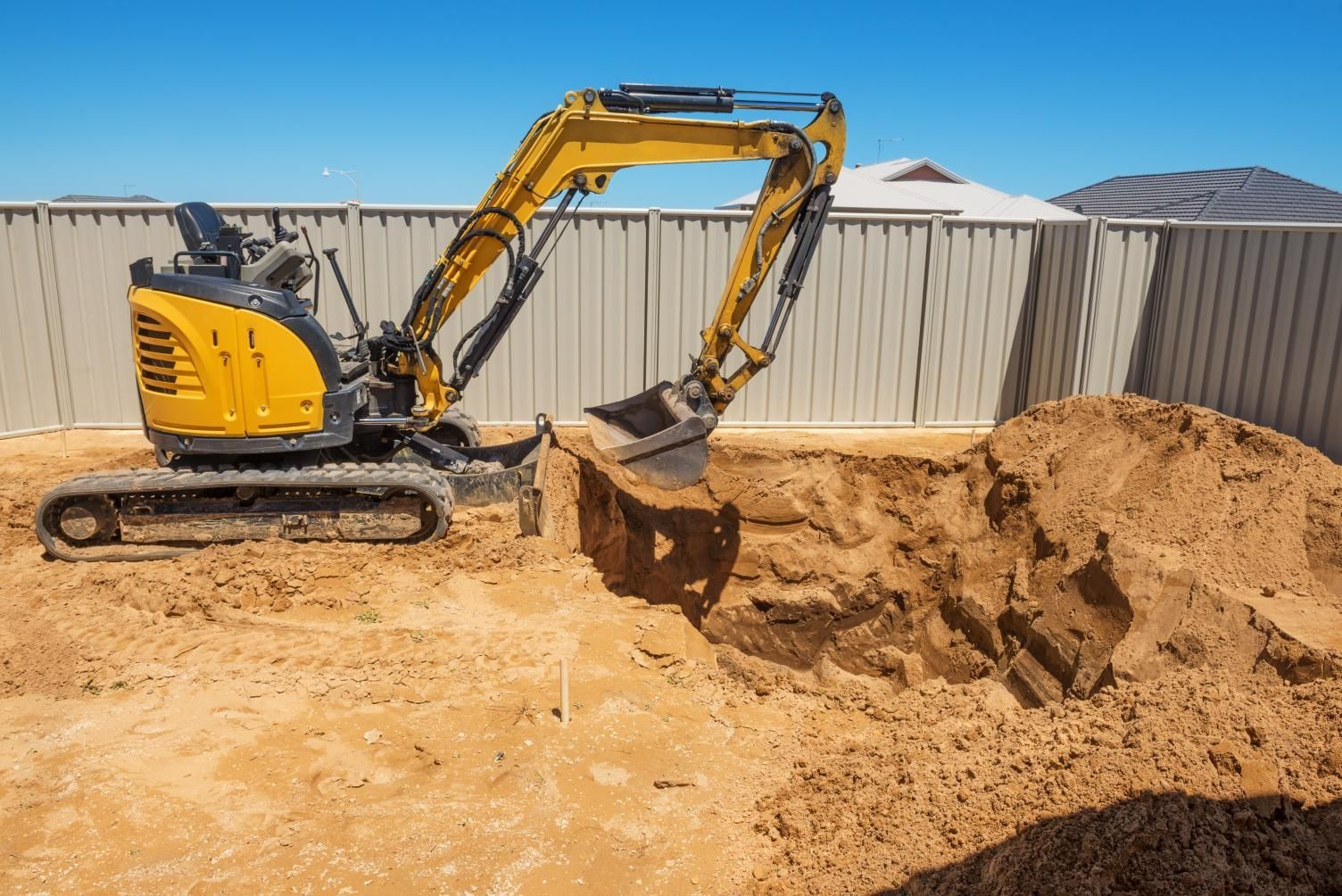 Yellow excavator digging a sandy trench beside a metal fence under a clear sky