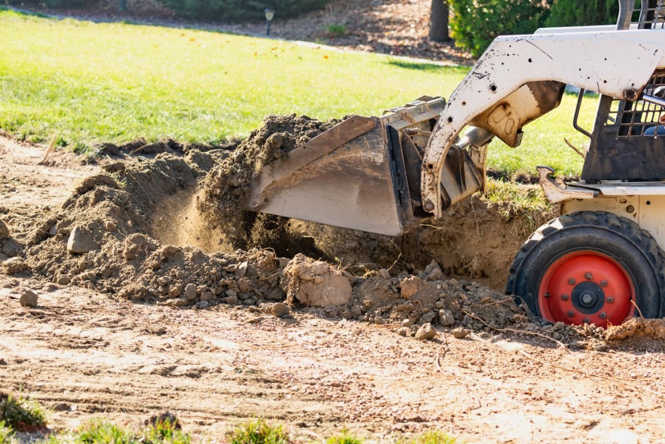 Backhoe digging dirt beside a grassy yard, with soil piled around the bucket