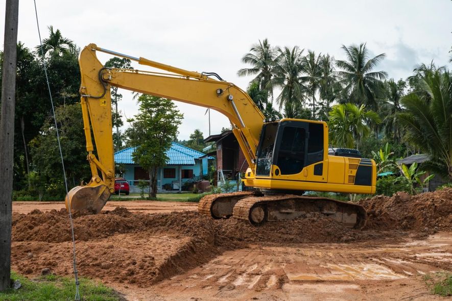 Yellow excavator digging dirt at a construction site with palm trees and a blue-roofed house in the background