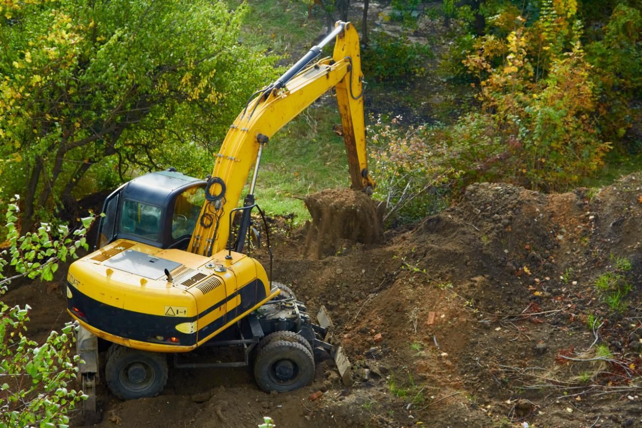 Yellow excavator digging earth on a wooded hillside, with dirt piled beside it.