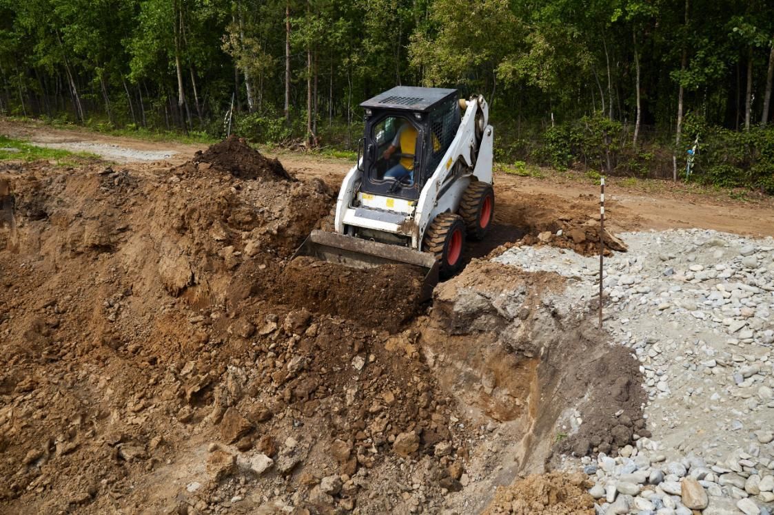 Skid steer excavator digging a dirt embankment beside a rocky path in a wooded area