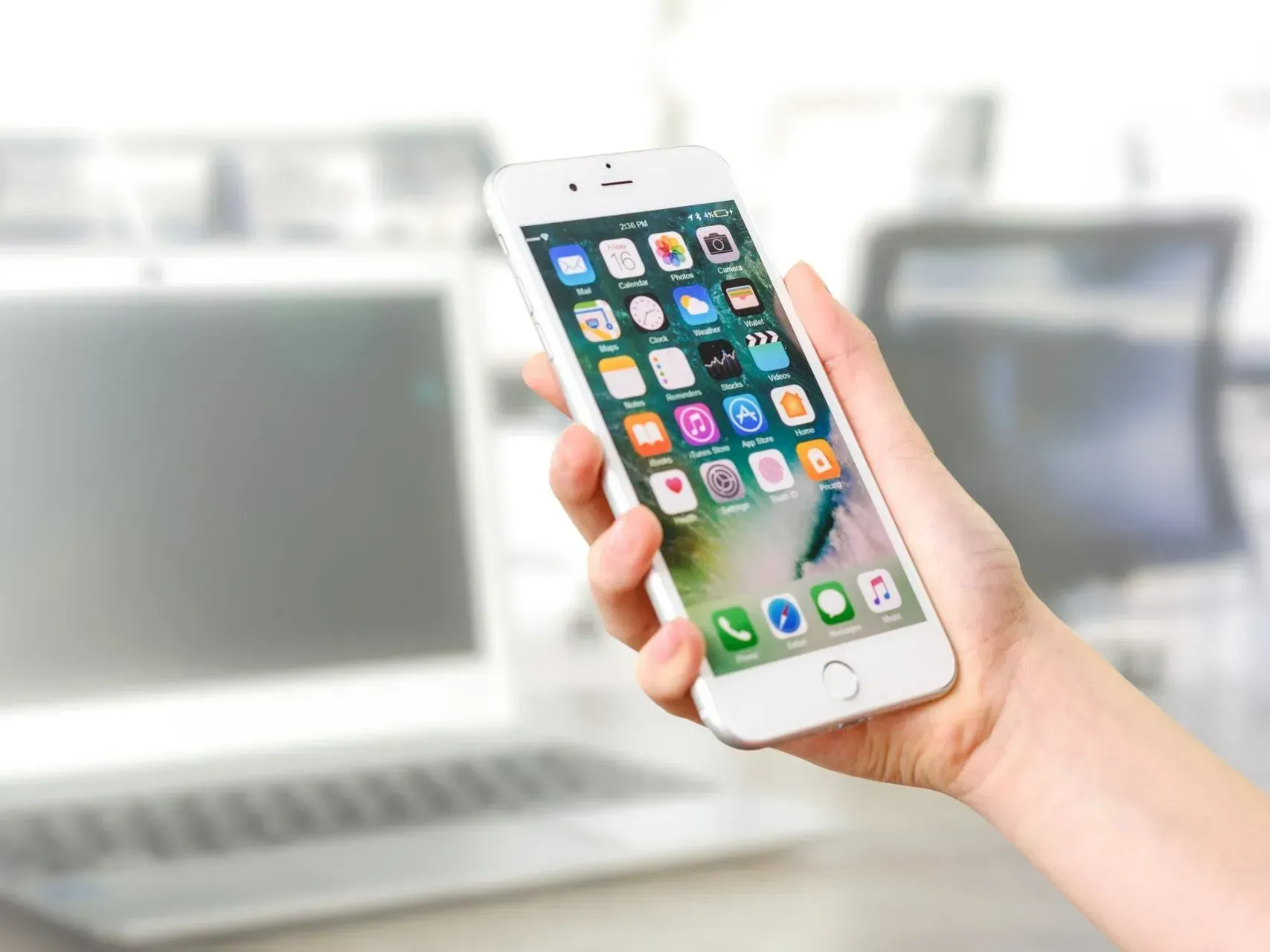 Hand holding a smartphone with app icons, in front of a blurred laptop on a desk.