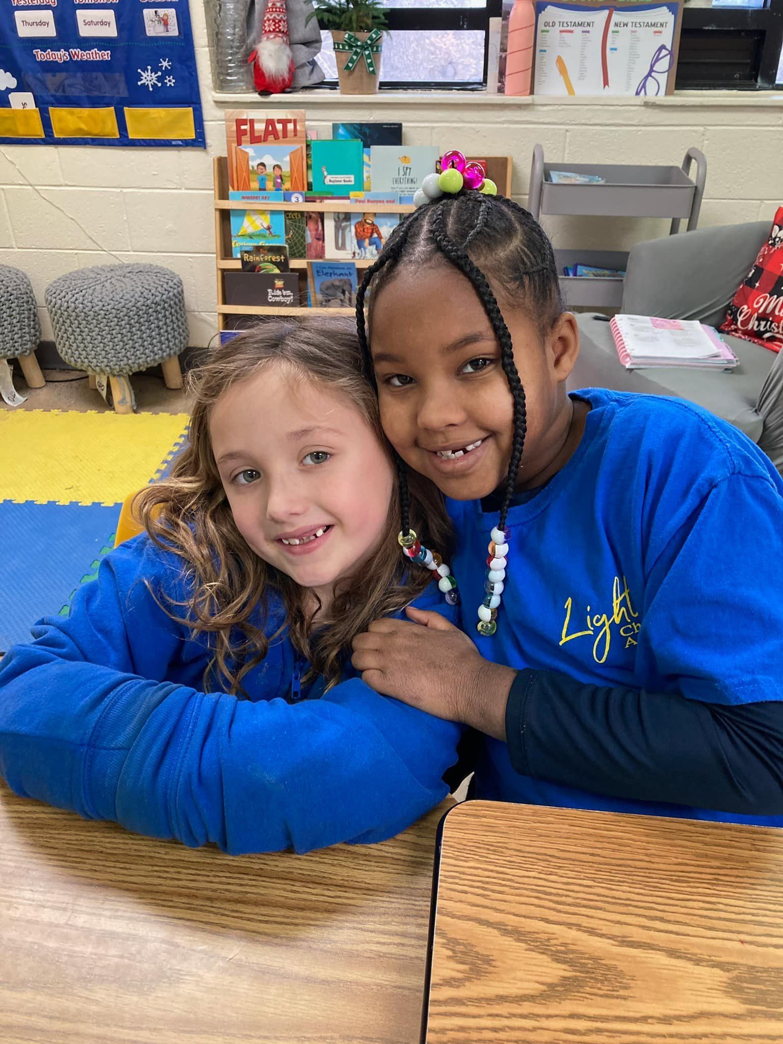 Two children in blue shirts smiling and hugging at a classroom table.